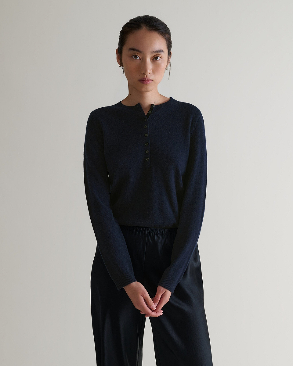 Image 4 of 6 showing a woman wearing Rise & Fall's indigo cashmere merino henley jumper with black silk trousers. 