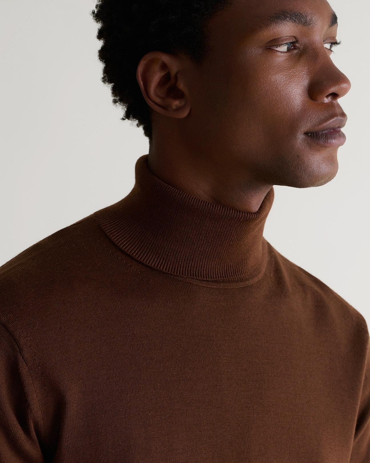 Man wears fine merino roll neck jumper in chestnut brown.