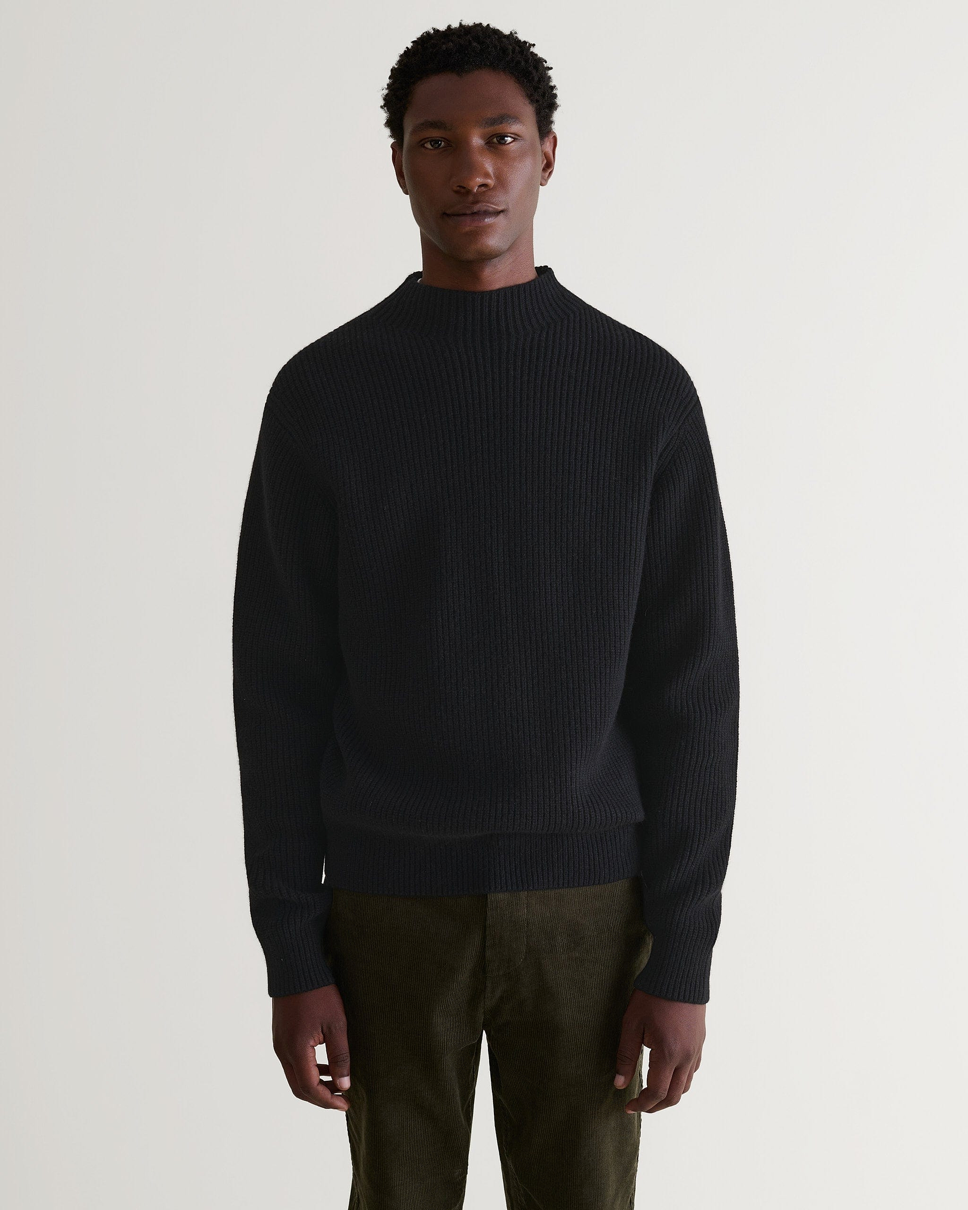 Front view of man wearing Rise & Fall's black cashmere merino stand neck ribbed sweater with dark green corduroy trousers.