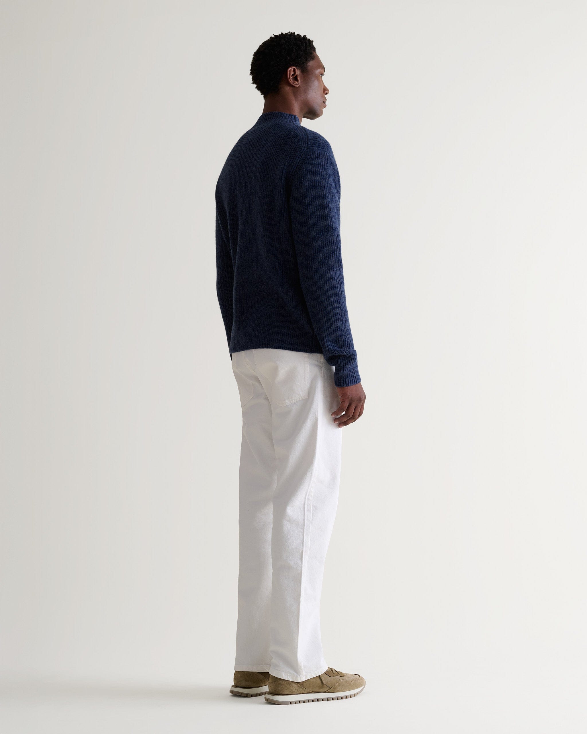 Side view of man wearing Rise & Fall blue cashmere merino stand neck jumper with white trousers and beige suede trainers.
