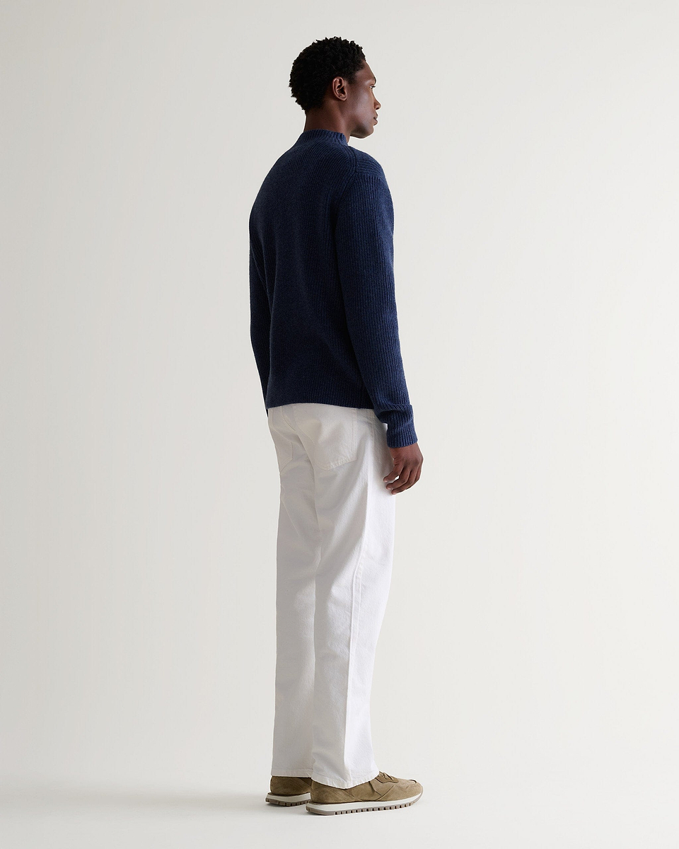Side view of man wearing Rise & Fall blue cashmere merino stand neck jumper with white trousers and beige suede trainers.