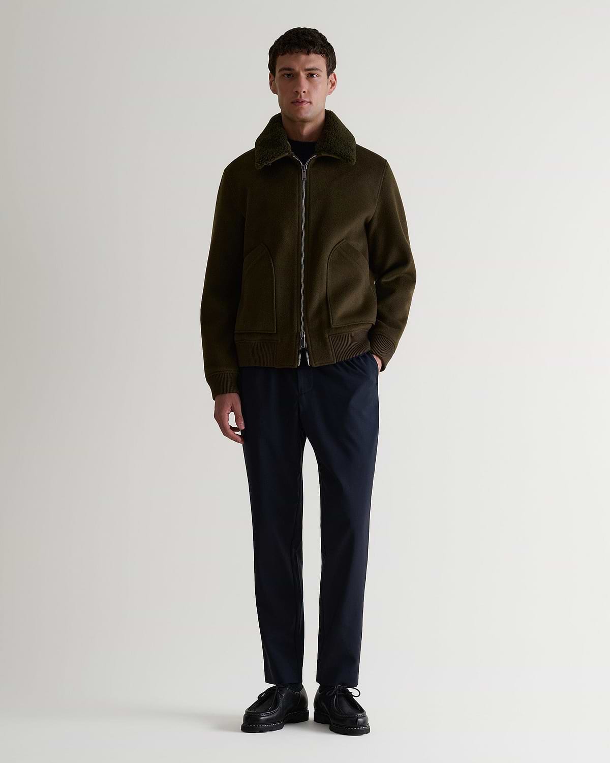 Full length view of man wearing Rise & Fall's olive wool bomber jacket with shearling collar. Matched with black trousers.