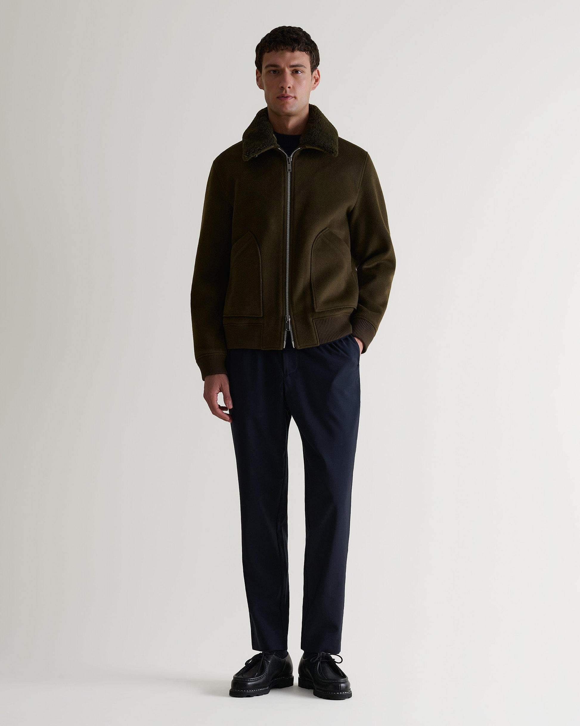 Full length view of man wearing Rise & Fall's olive wool bomber jacket with shearling collar. Matched with black trousers. 