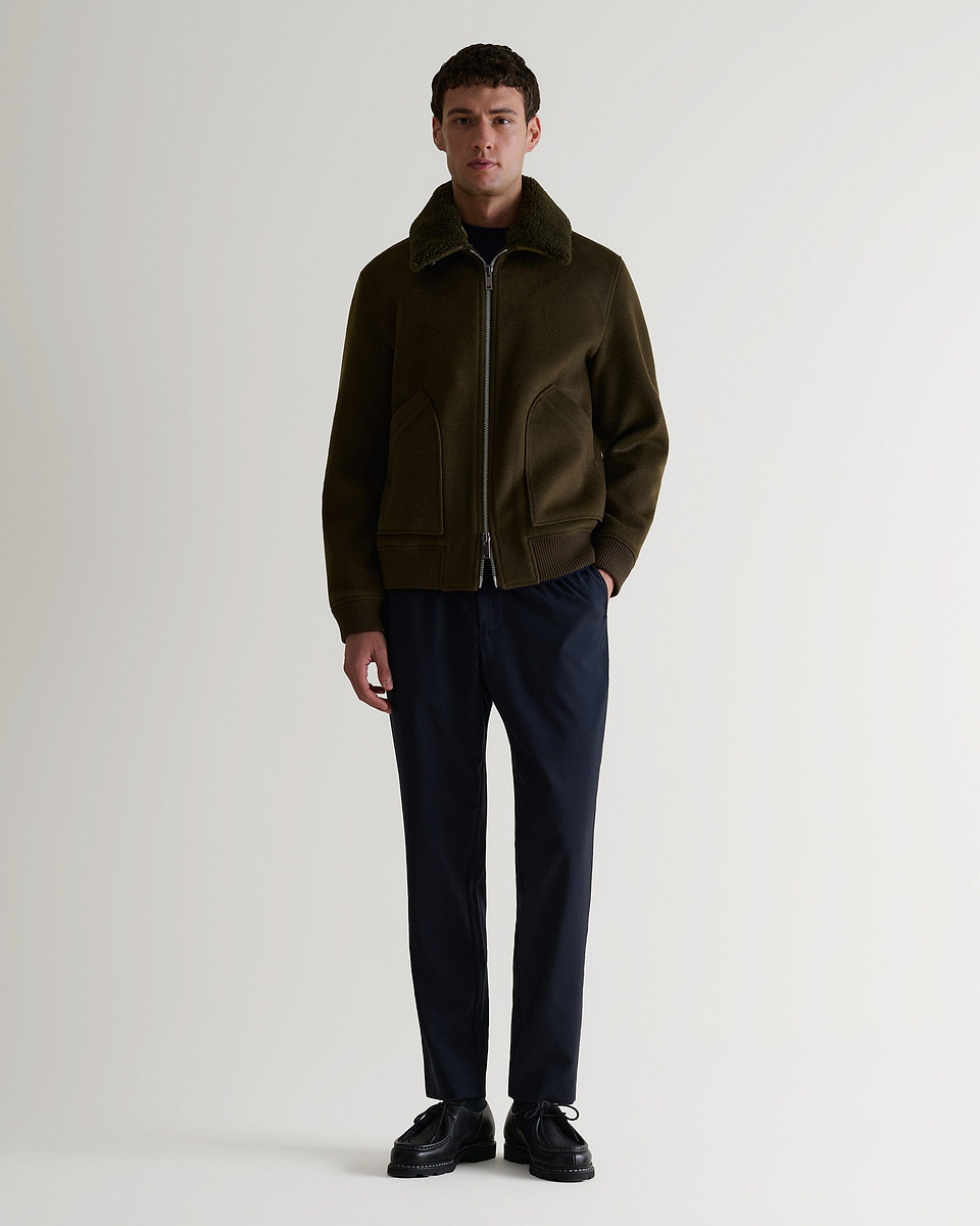 Full length view of man wearing Rise & Fall's olive wool bomber jacket with shearling collar. Matched with black trousers. 