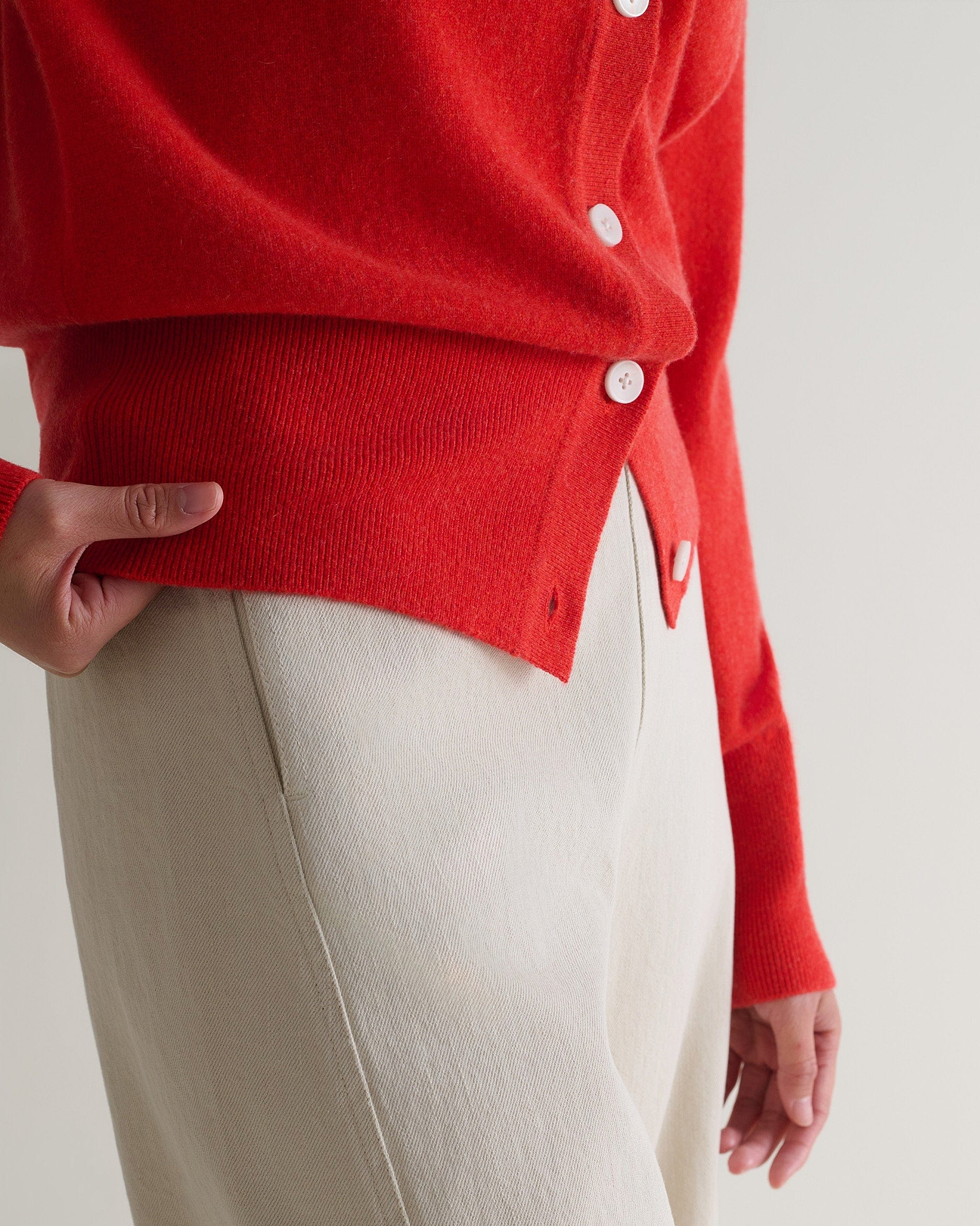 A close up of Rise & Fall's poppy red fine cashmere blouson cardigan worn with cream trousers.