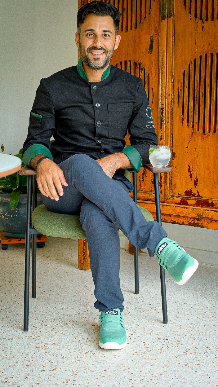 Man in a chef's coat sitting with a drink, wearing green sneakers.