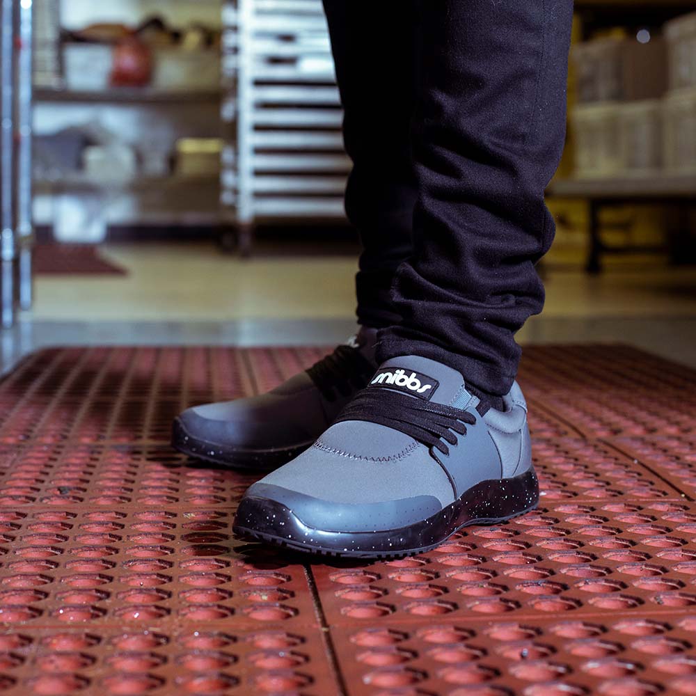 Person wearing gray sneakers with black laces, standing on a patterned mat.