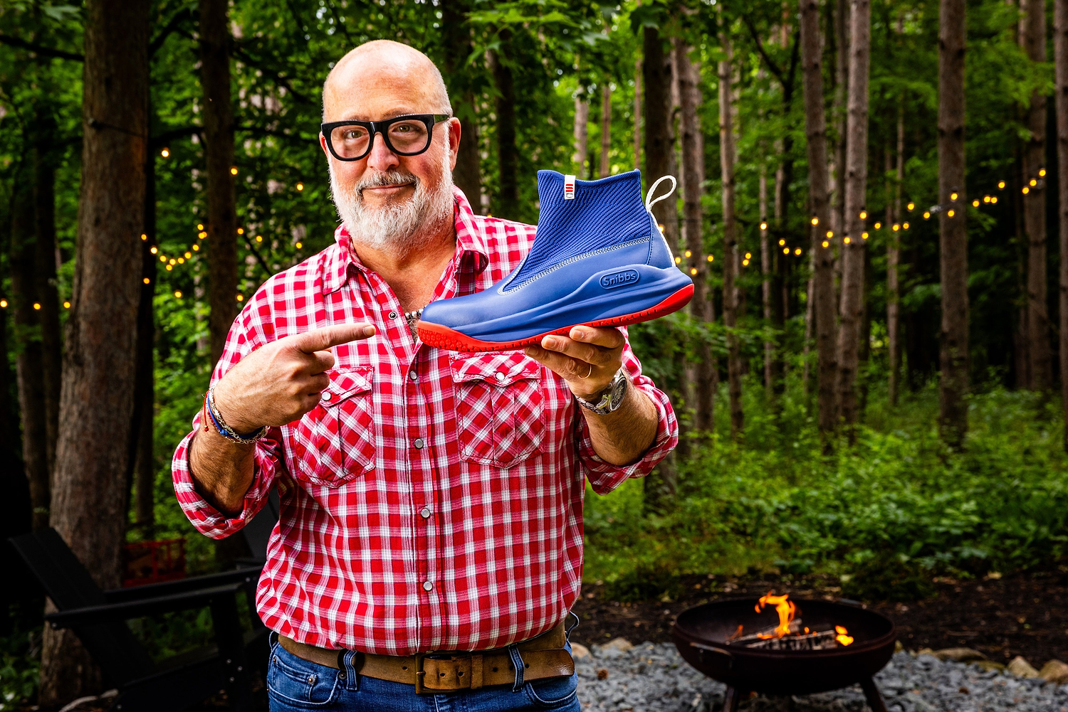 Andrew Zimmern holding a blue sneaker outdoors with trees and string lights in the background
