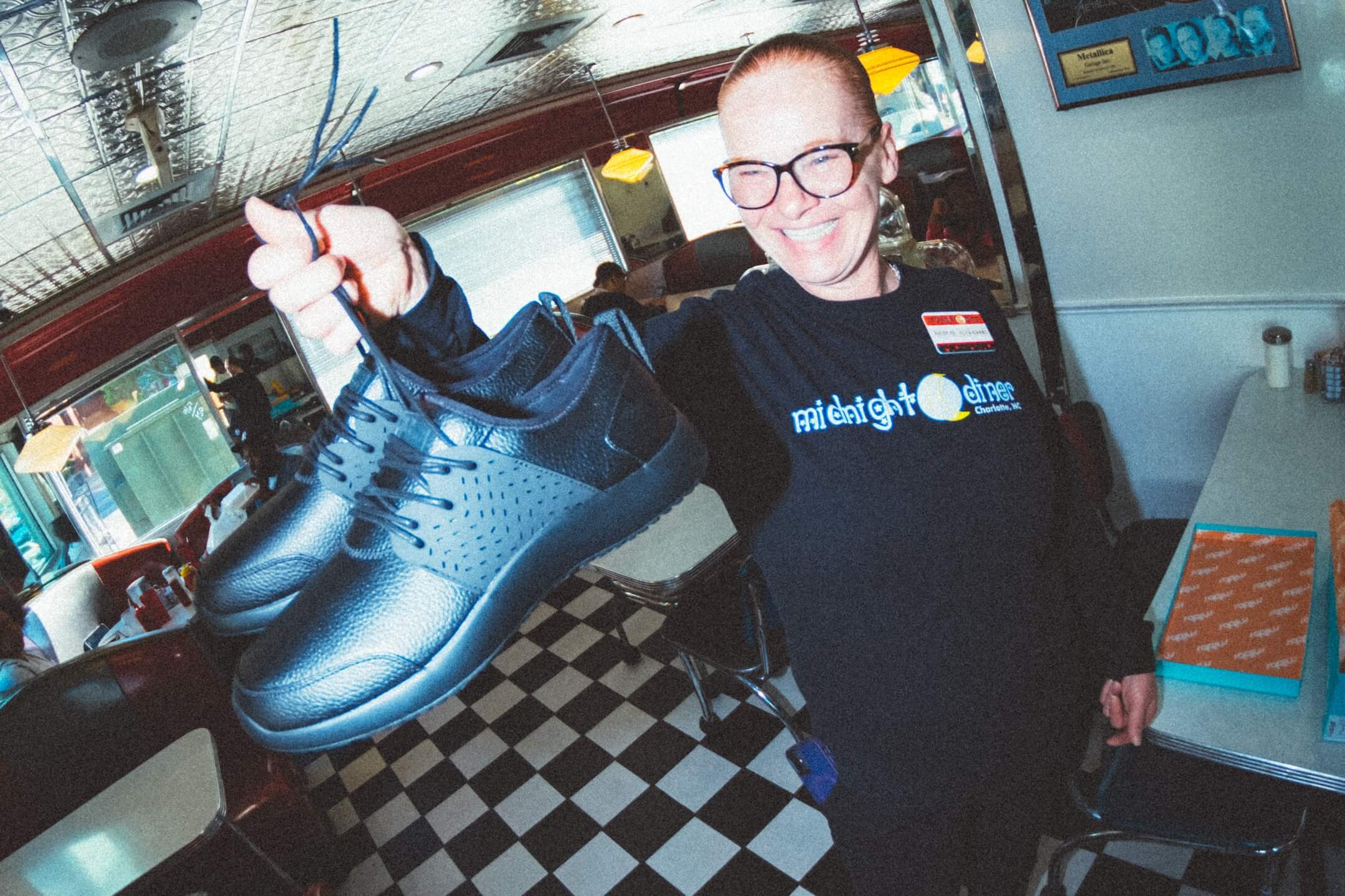 A smiling person in glasses holds up a pair of new black shoes inside a retro diner with classic black-and-white checkered flooring and a vibrant atmosphere.