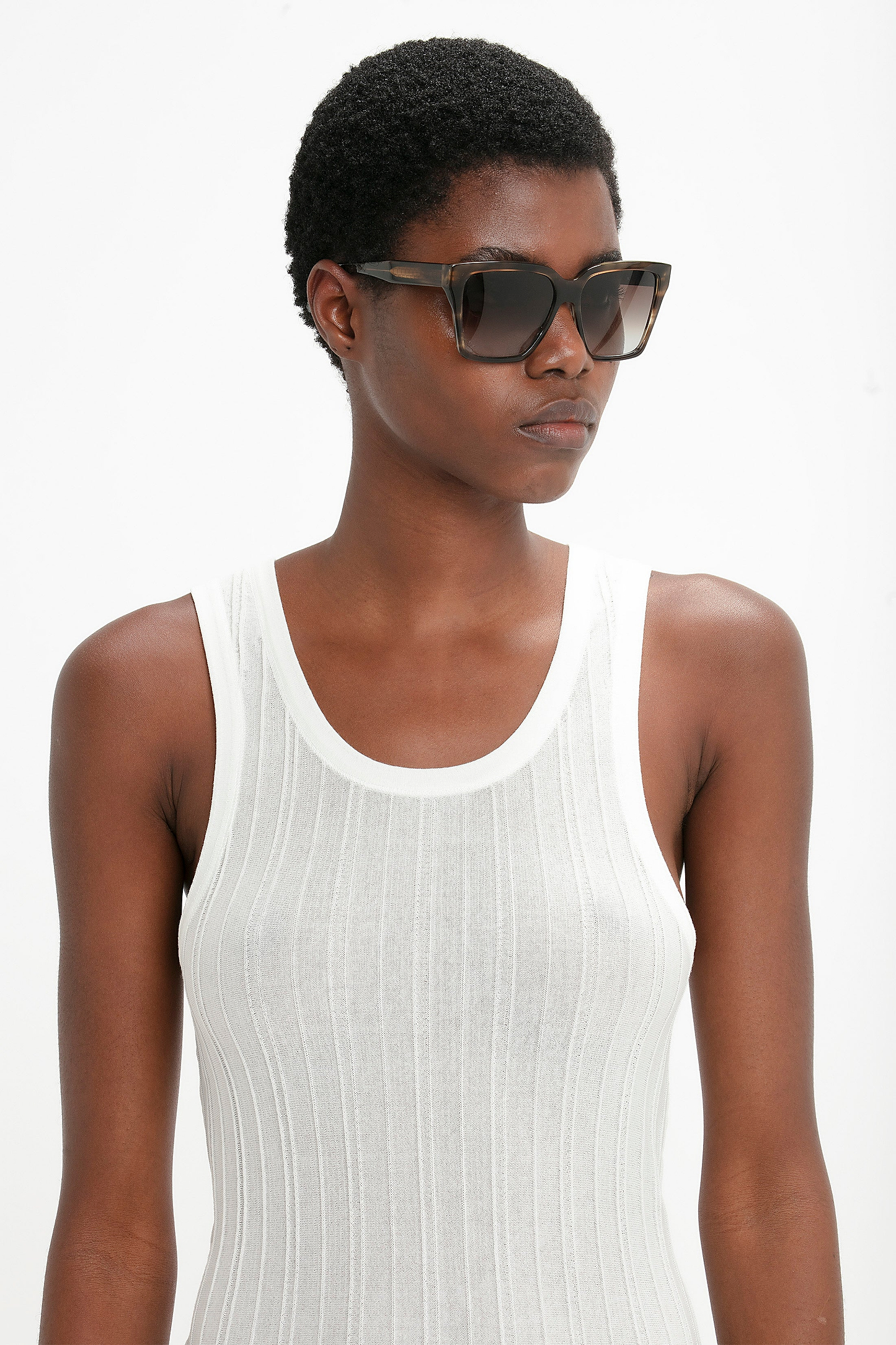 Person with short hair wearing Victoria Beckham Soft Square Frame Sunglasses In Striped Khaki Havana and a white sleeveless ribbed top, standing against a plain background.