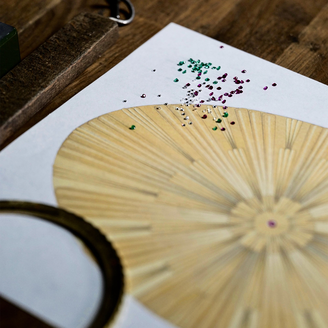 Concept sketch and gemstone layout for the Fabergé Centenary Egg, featuring radial guilloché-inspired design details with scattered coloured gemstones on a jeweller’s workbench.