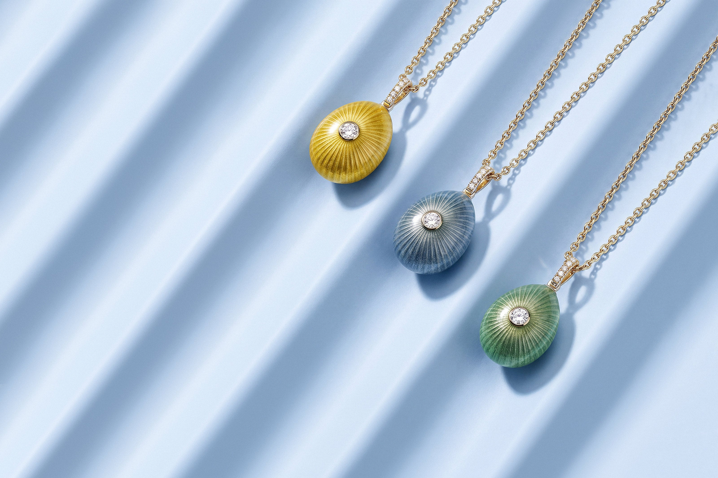Fabergé Sunburst Egg Pendants in yellow, blue and green enamel, each featuring a central diamond and intricate radiating sunburst detailing, displayed on gold chains against a soft blue backdrop.