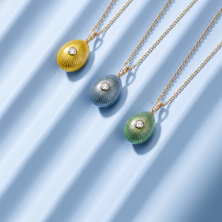 Mobile Banner image of Fabergé Sunburst Egg Pendants in yellow, blue and green enamel, each featuring a central diamond and intricate radiating sunburst detailing, displayed on gold chains against a soft blue backdrop.