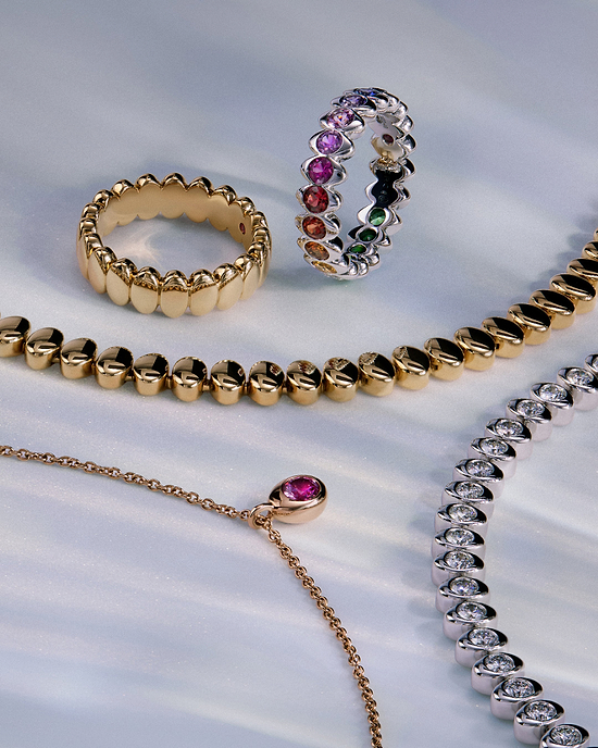 Flat lay of Fabergé Code gold and diamond jewellery including gemstone eternity rings, diamond bracelets, and a pink sapphire pendant necklace on a soft textured background