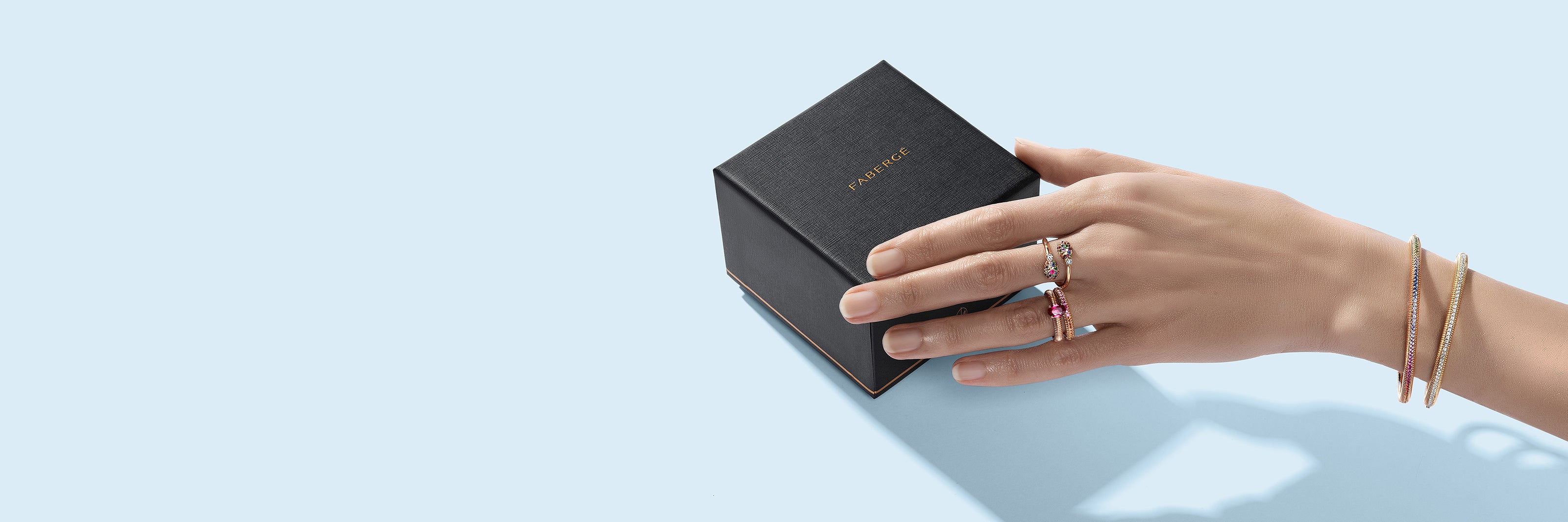 Hand with a selection of Fabergé rings next to a black jewelry box on a light blue background