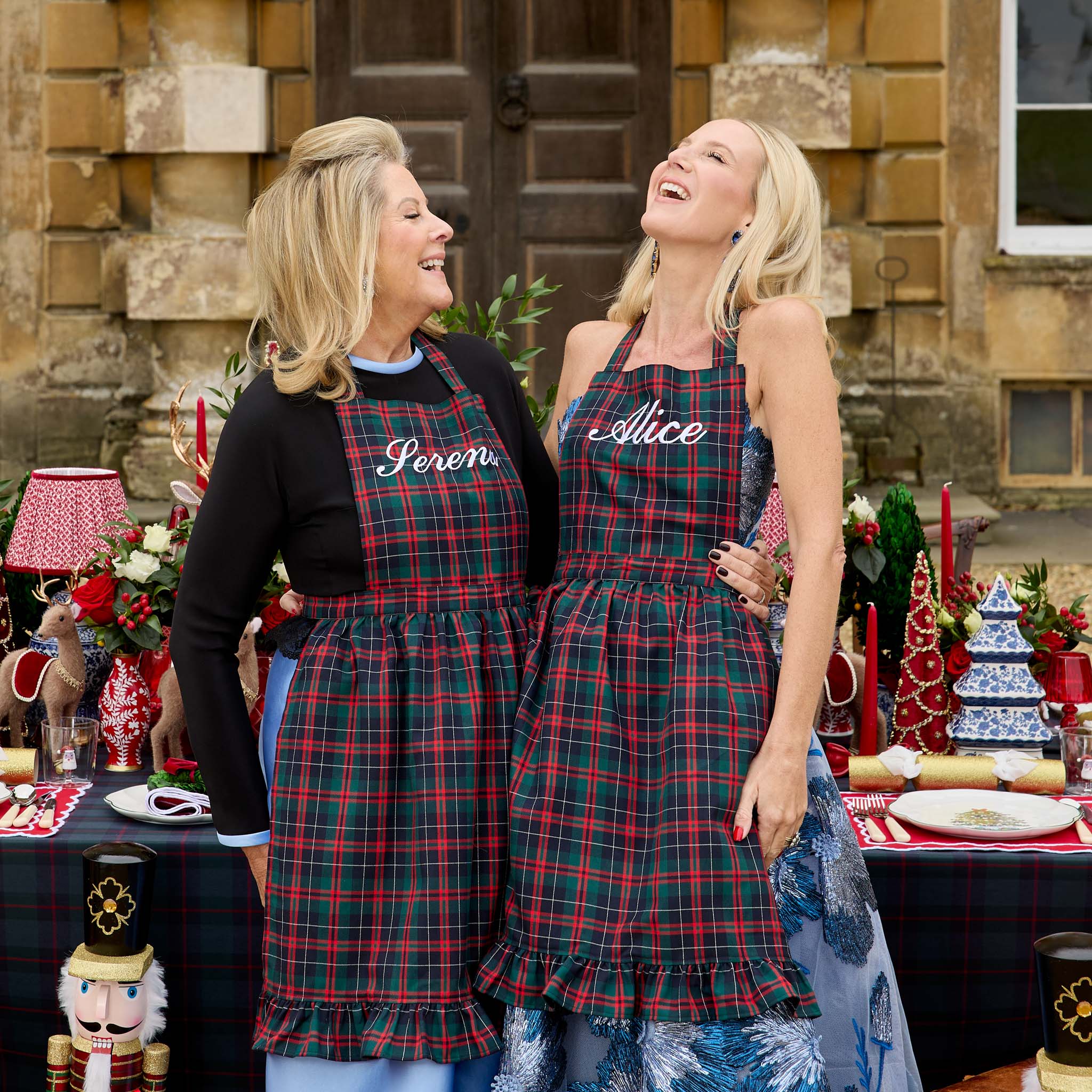 Two women wearing plaid aprons with names on them, standing in front of a decorated table.