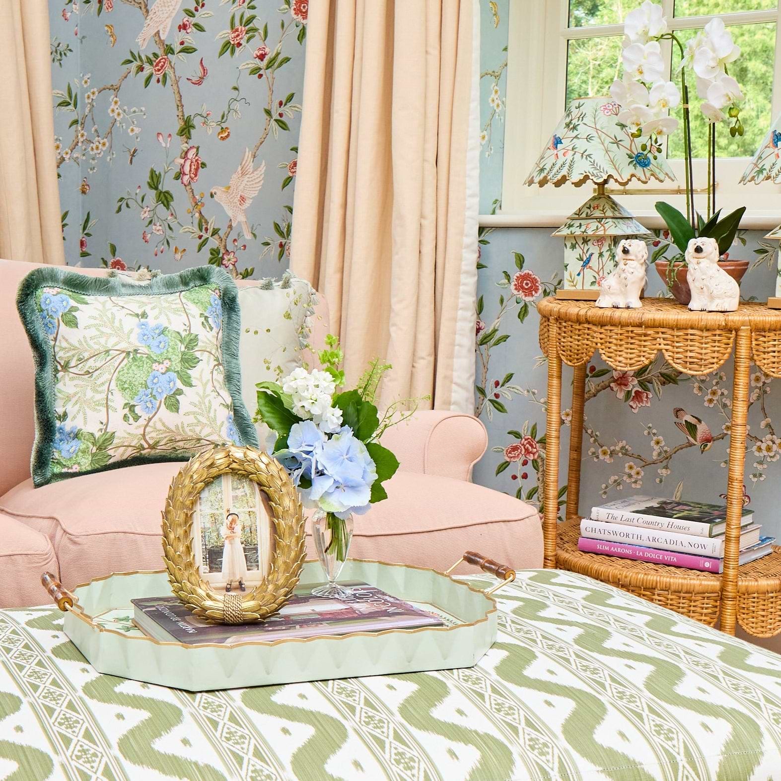A large upholstered storage ottoman with a green and cream geometric ikat pattern, styled in a bright, vintage-inspired living room with a pink sofa and floral wallpaper.