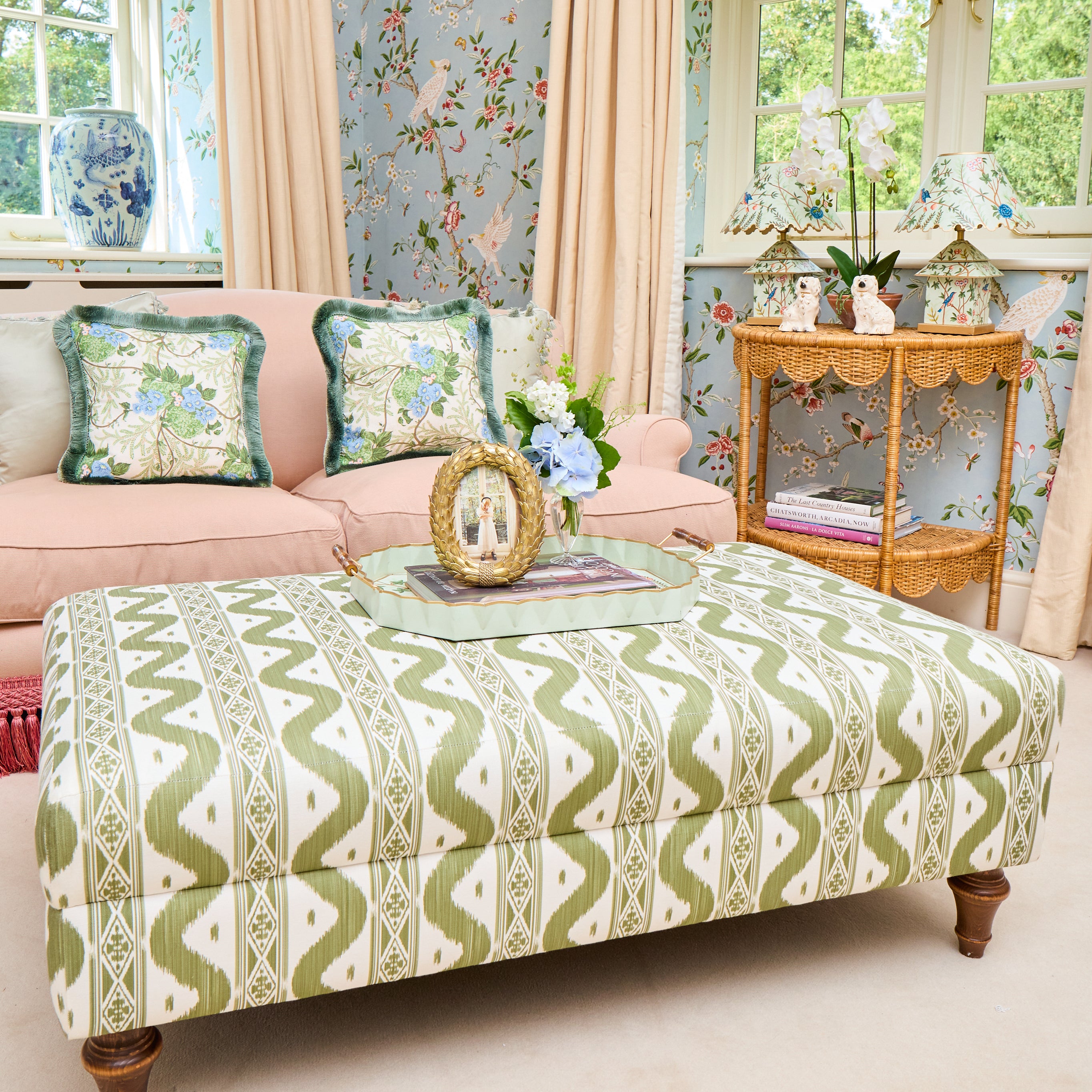 A large upholstered storage ottoman with a green and cream geometric ikat pattern, styled in a bright, vintage-inspired living room with a pink sofa and floral wallpaper.