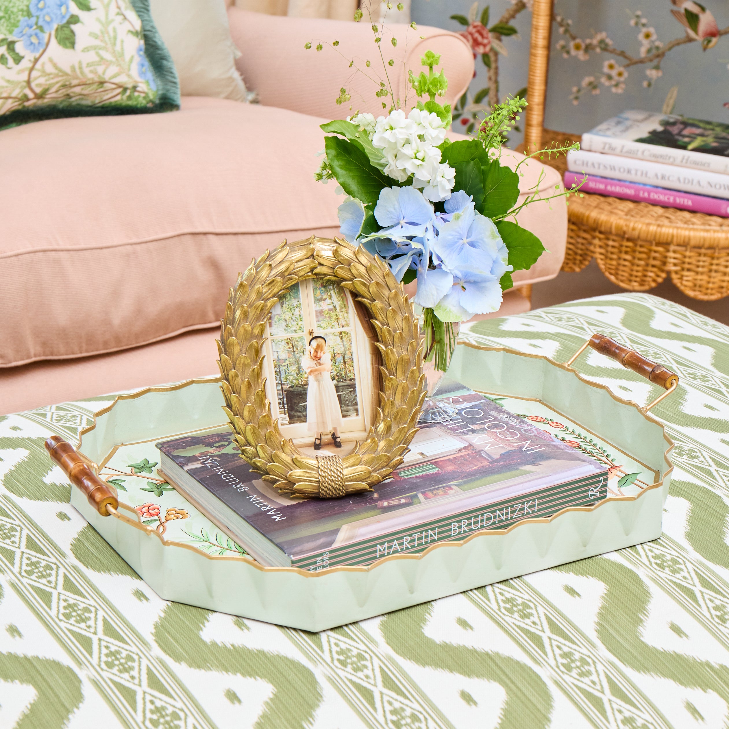 A decorative rectangular serving tray with wooden handles and a scalloped edge, shown in a styled living room setting. The tray is pale mint green with gold trim and floral detailing, holding a stack of magazines and a round woven gold frame containing a photo of a woman.