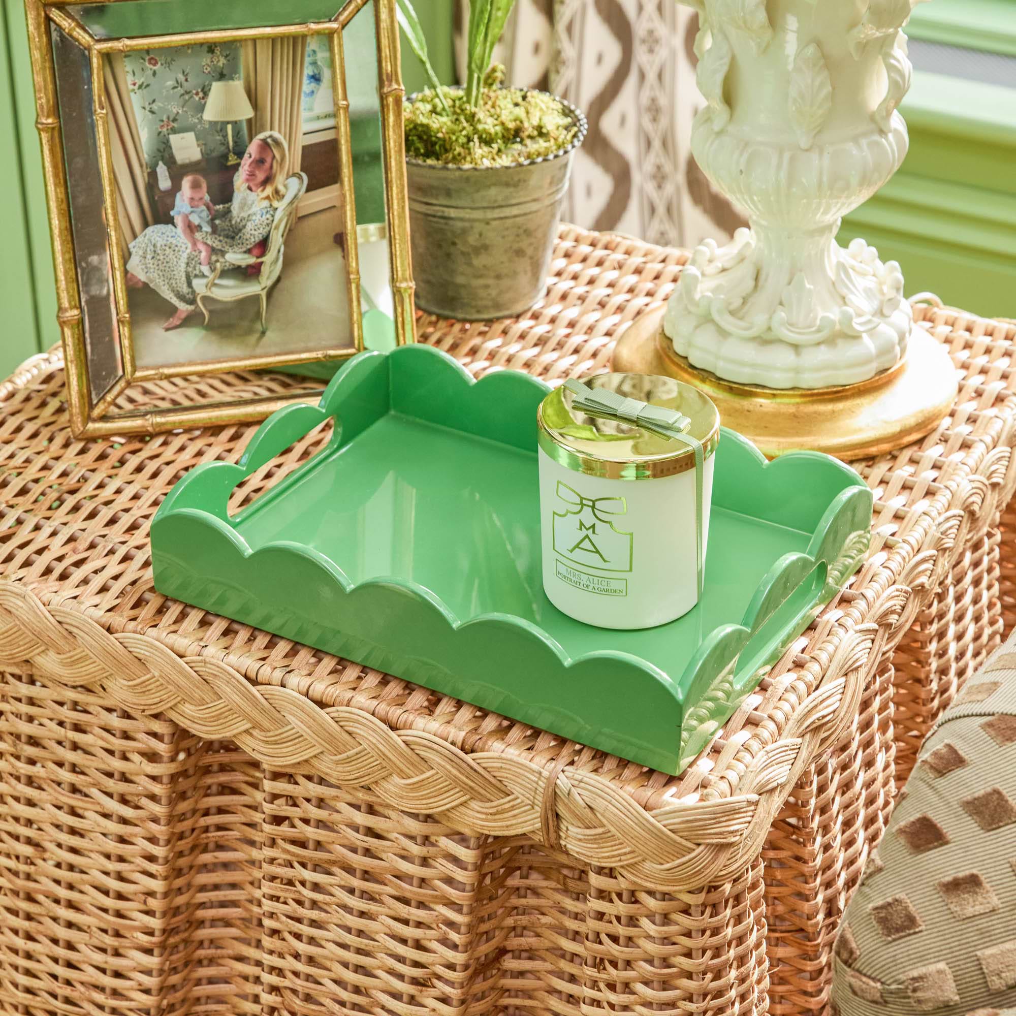 Green decorative tray with a candle on a wicker surface, surrounded by a photo frame, plant, and decorative urn.