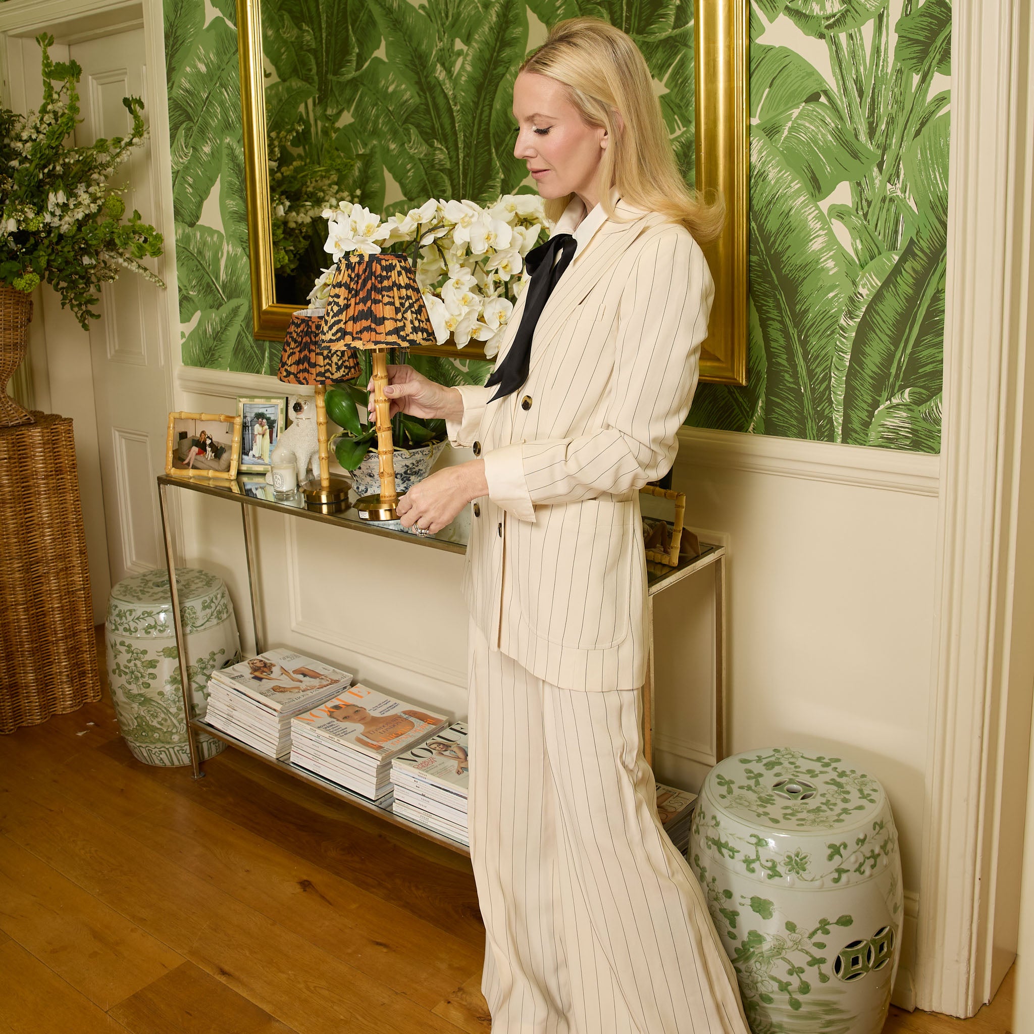 Woman in a white suit standing in a room with green leafy wallpaper and decorative items.