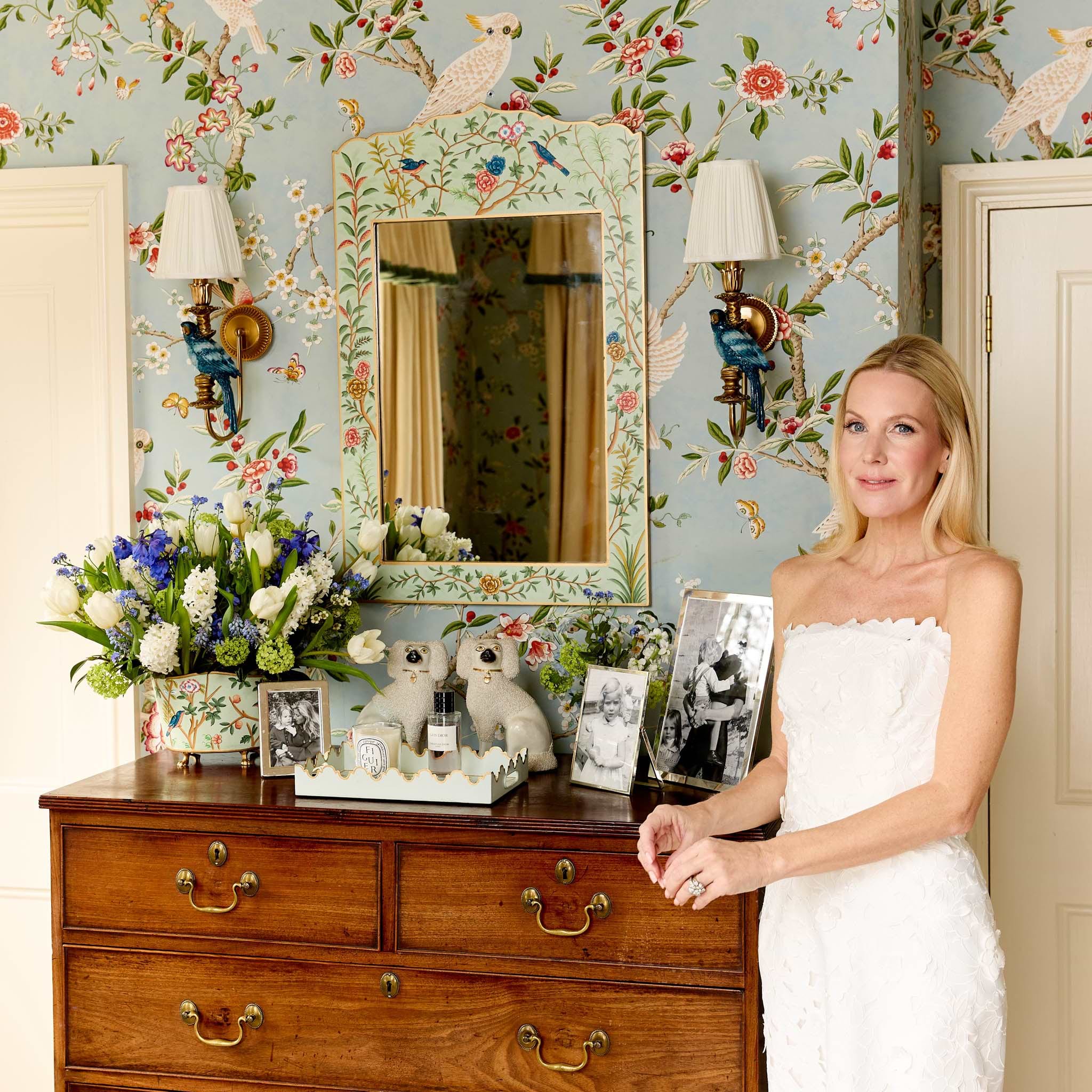 Woman in a white dress standing in a room with floral wallpaper, a wooden dresser, and decorative items.