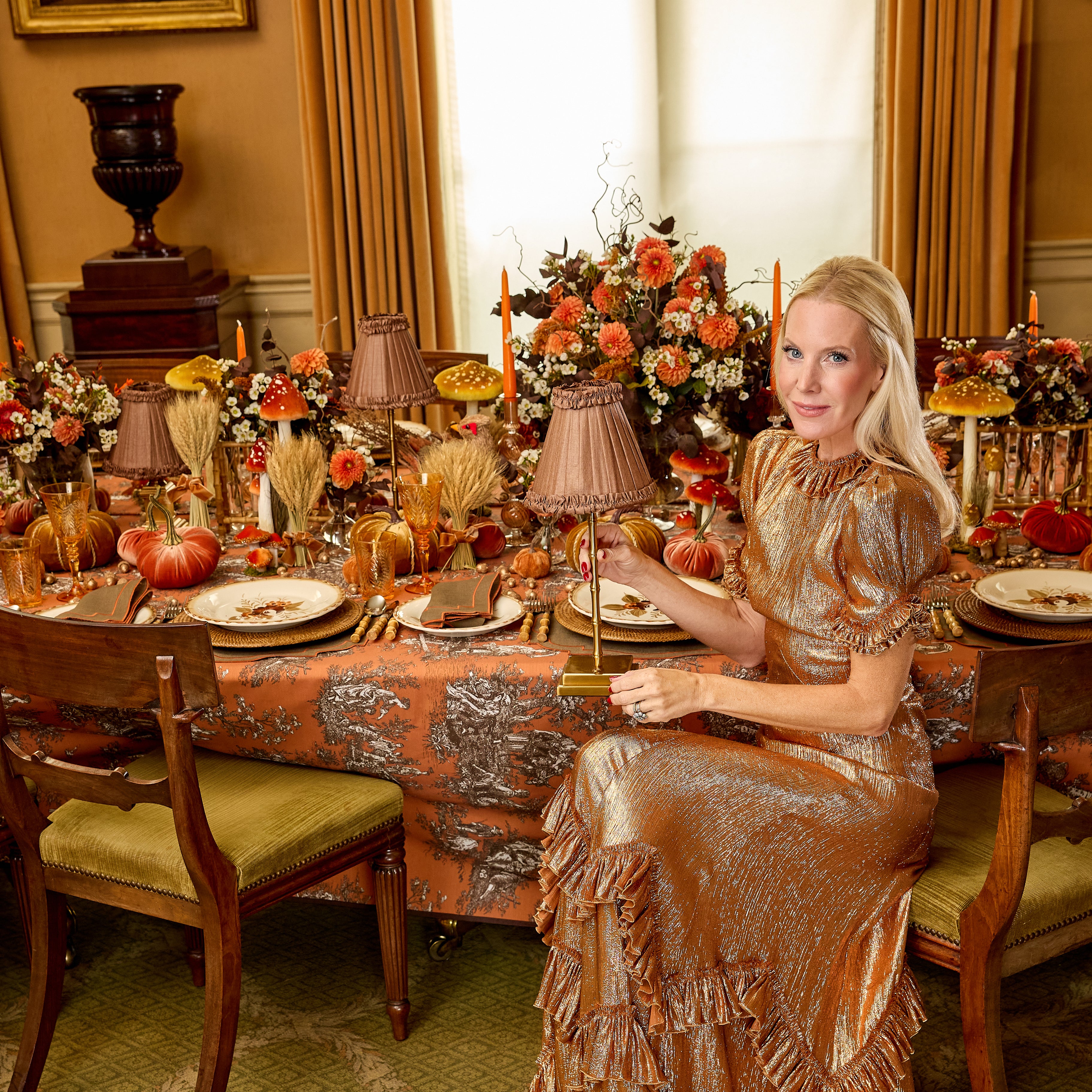 A woman in a shimmering bronze metallic dress sits at a decorated autumn table with pumpkins, floral arrangements, candles, and patterned linens in a warm, elegant dining room.