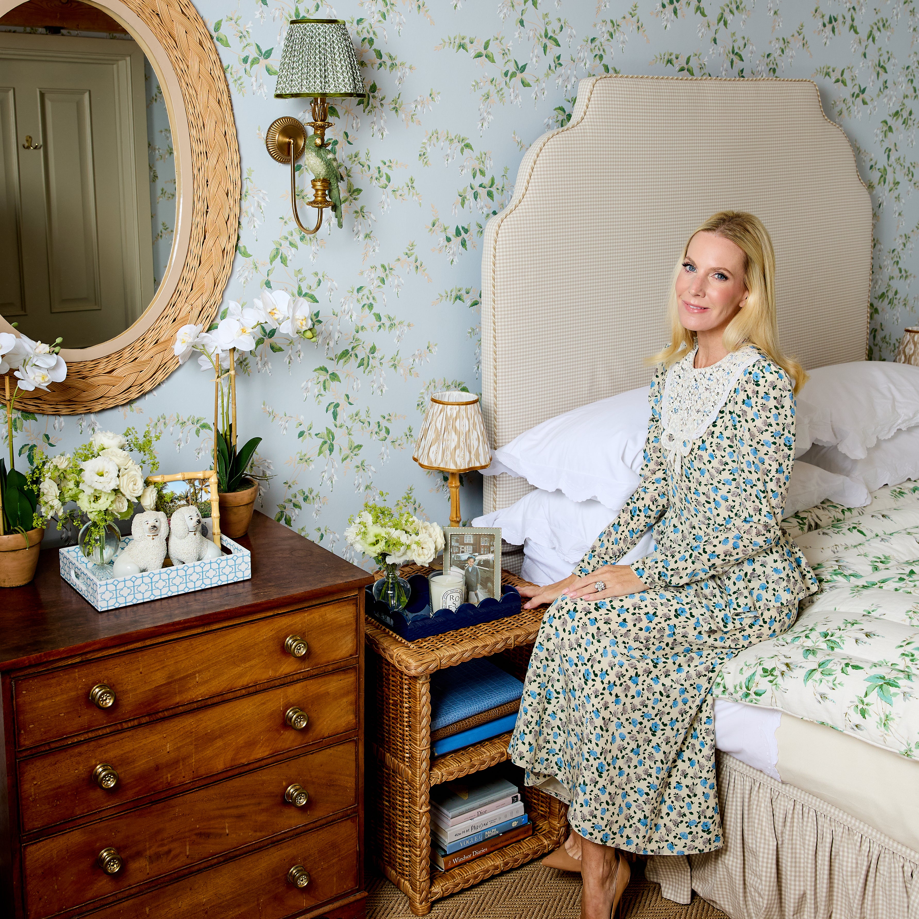 A woman sits on a bed wearing a long-sleeved floral midi dress with a white lace collar, in a beautifully decorated bedroom with green and white floral wallpaper, a wooden dresser, and a wicker bedside table.