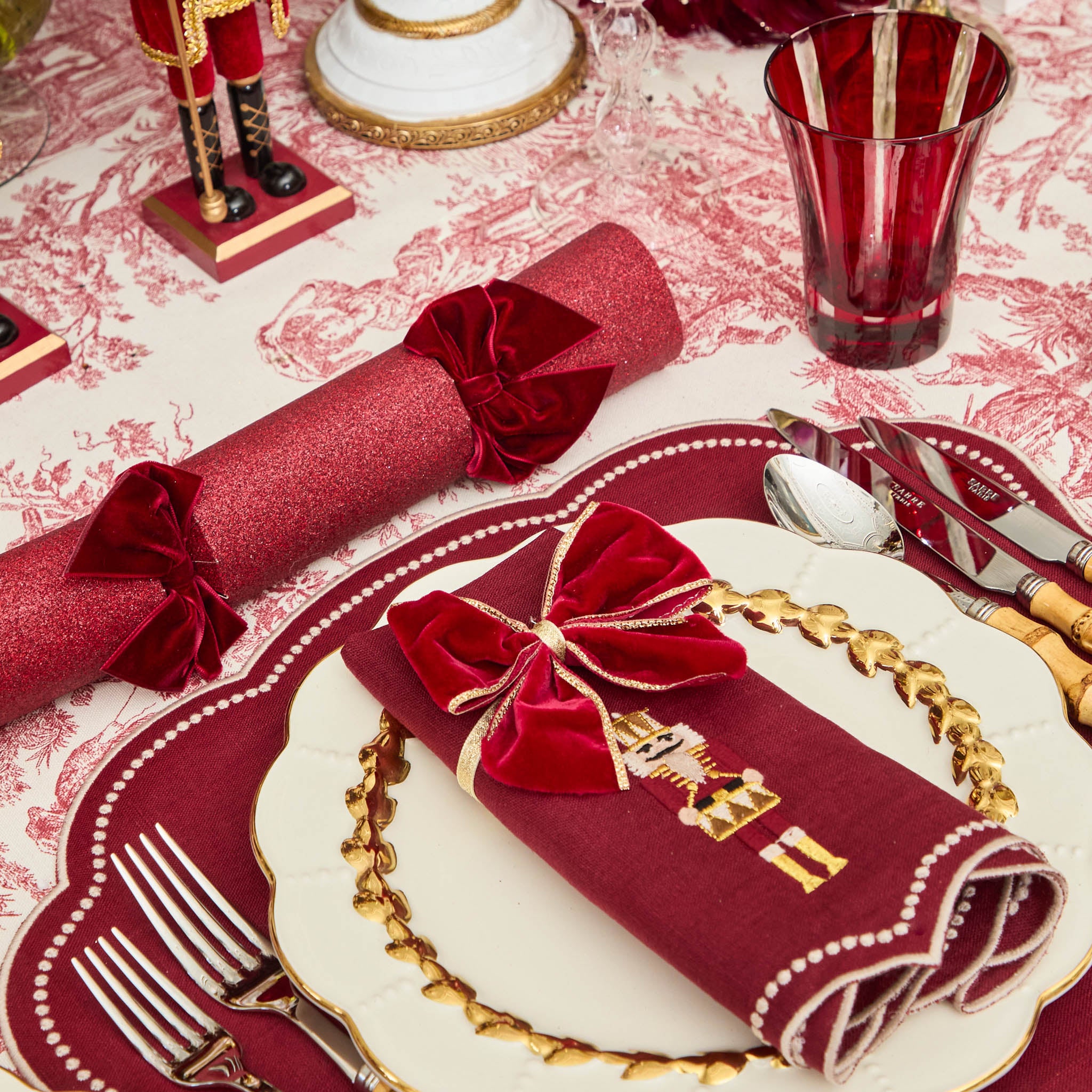 Decorative table setting with red napkins, gold cutlery, and a red glass on a patterned tablecloth.