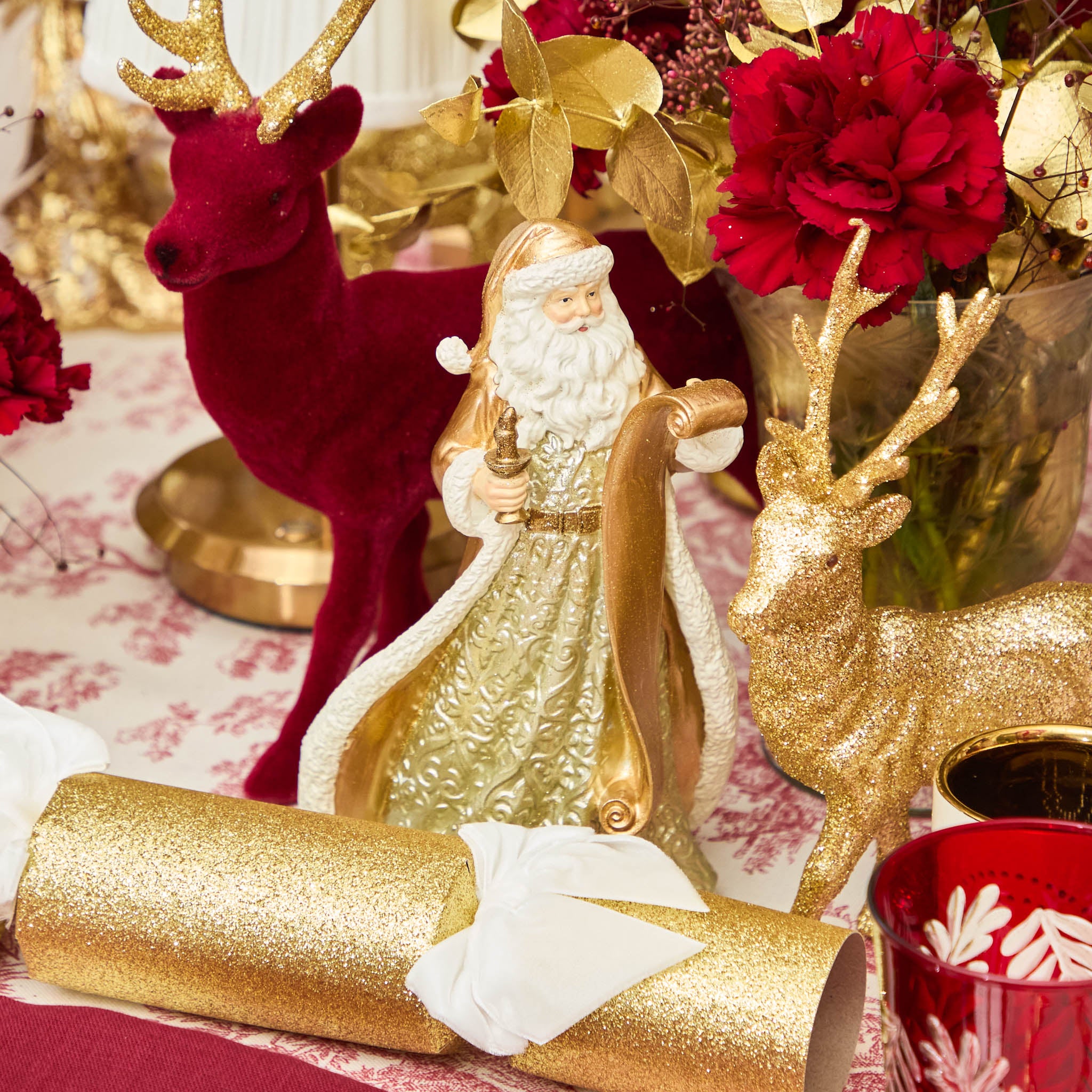 Decorative Christmas scene with gold Santa figure, red and gold reindeer, and red flowers on a patterned tablecloth.