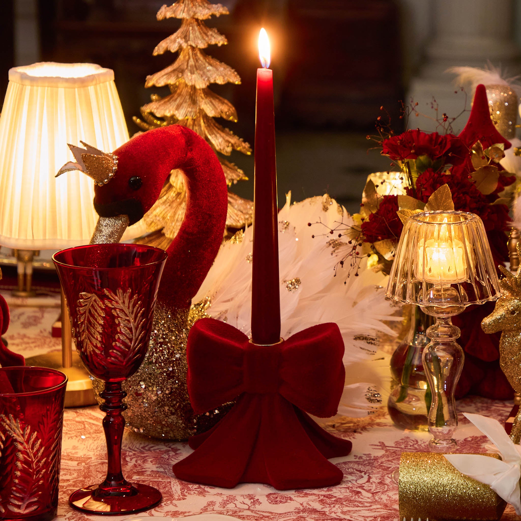 Decorative table setting with red candle, swan-shaped holder, and red goblet on a patterned tablecloth.