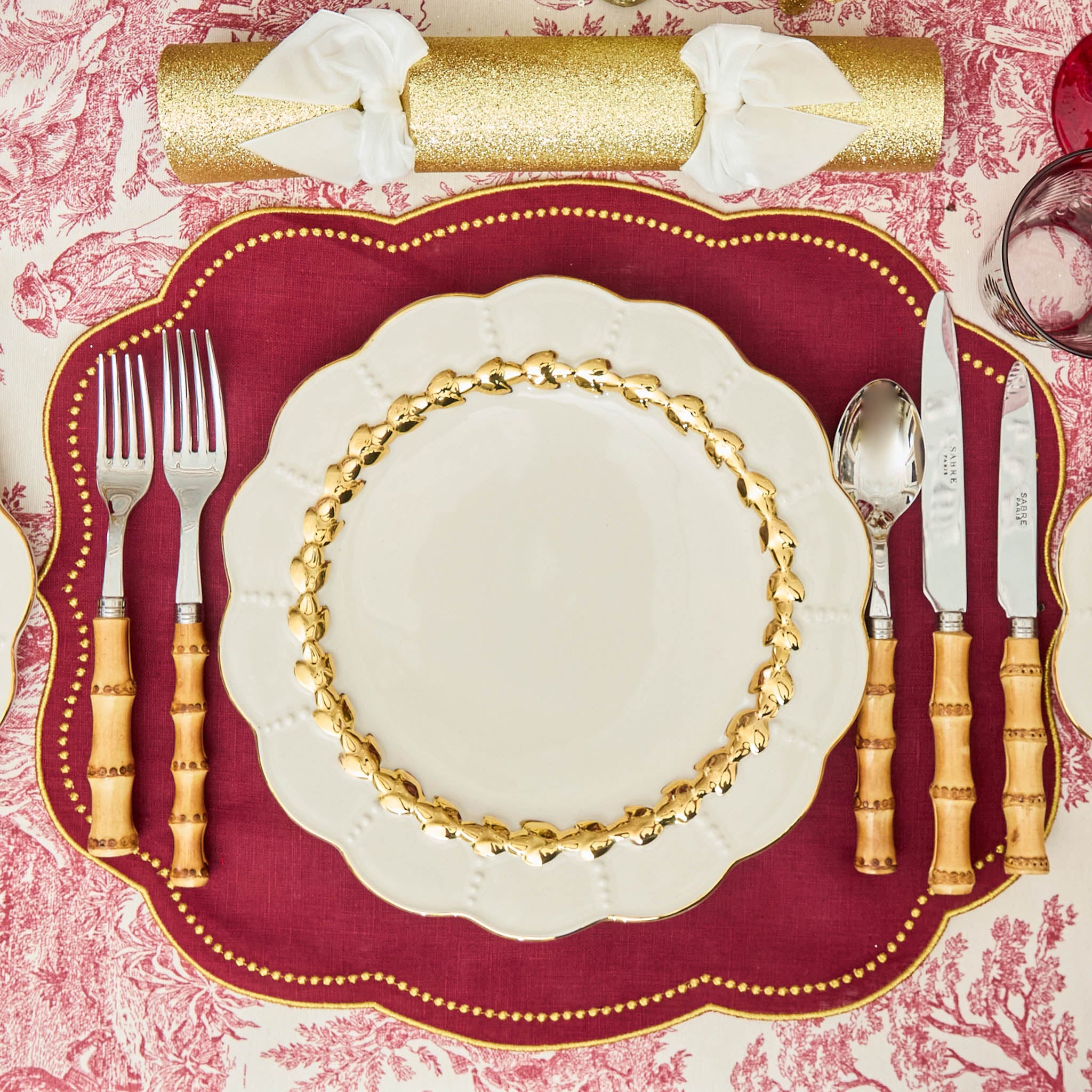 Decorative table setting with gold-rimmed plates, cutlery, and a red placemat on a patterned tablecloth.