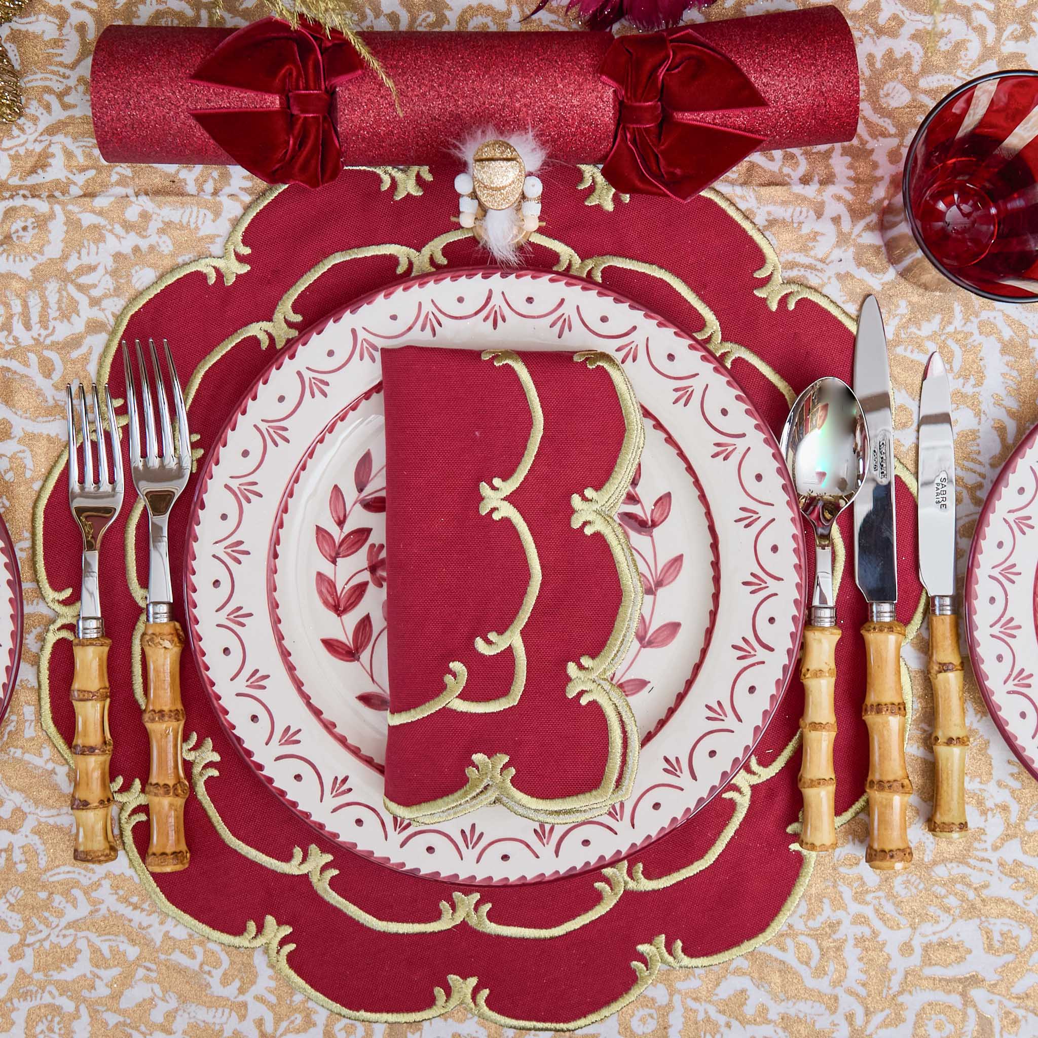 Decorative Christmas table setting with red and gold plates, napkins, and cutlery on a patterned tablecloth.