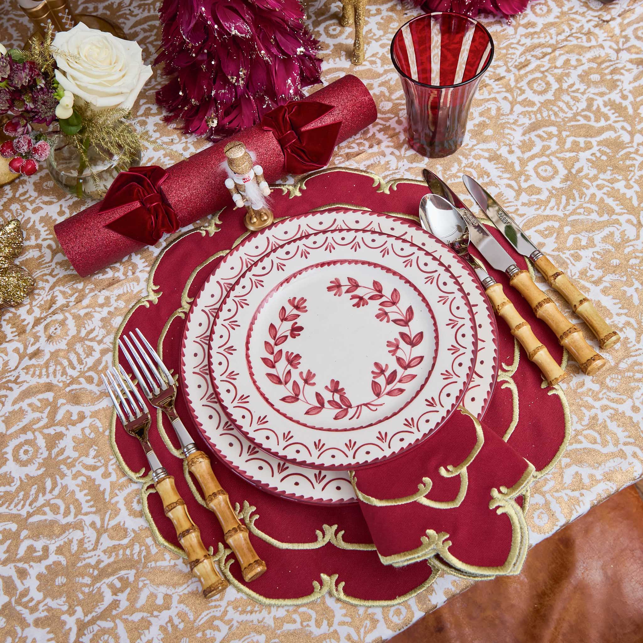 Decorative table setting with red and gold plate, cutlery, and napkins on a patterned tablecloth.