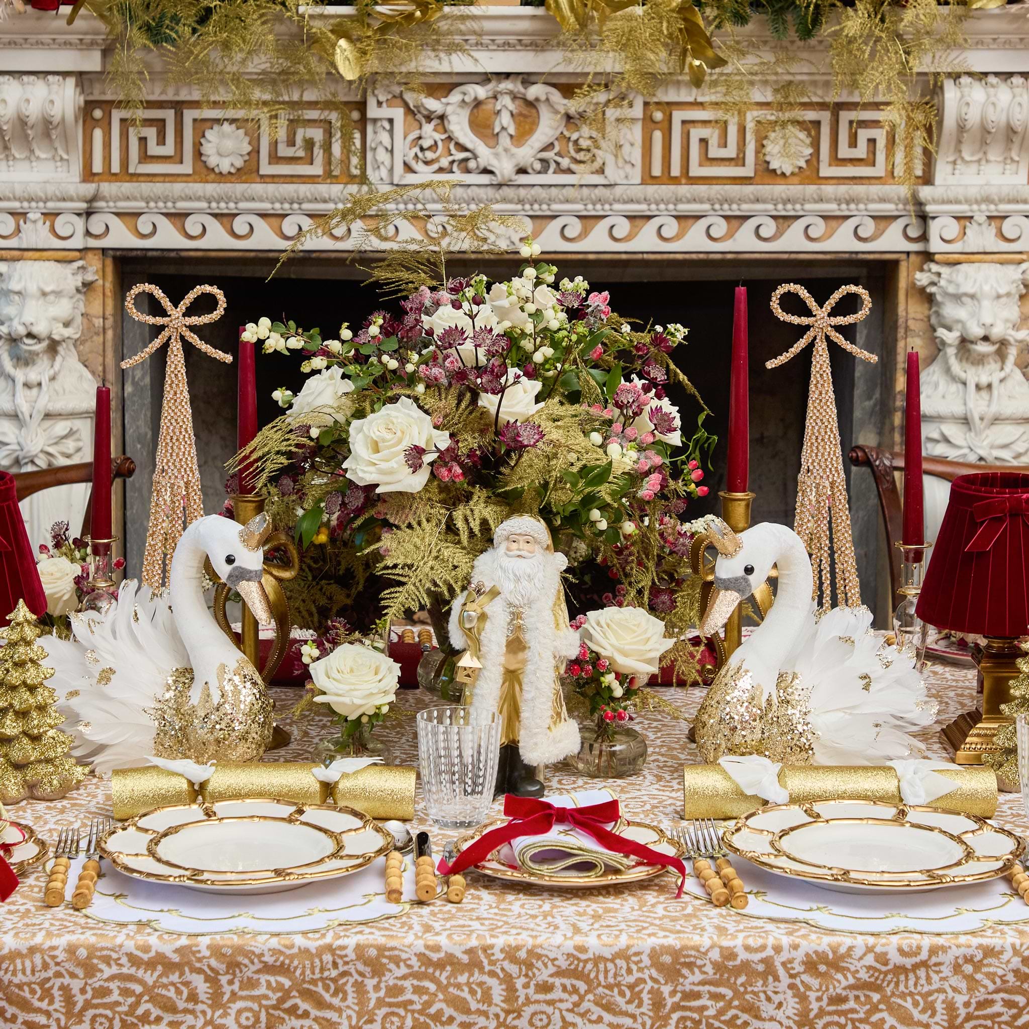 Decorative Christmas table setting with swans, Santa figure, and floral arrangement in front of a fireplace.