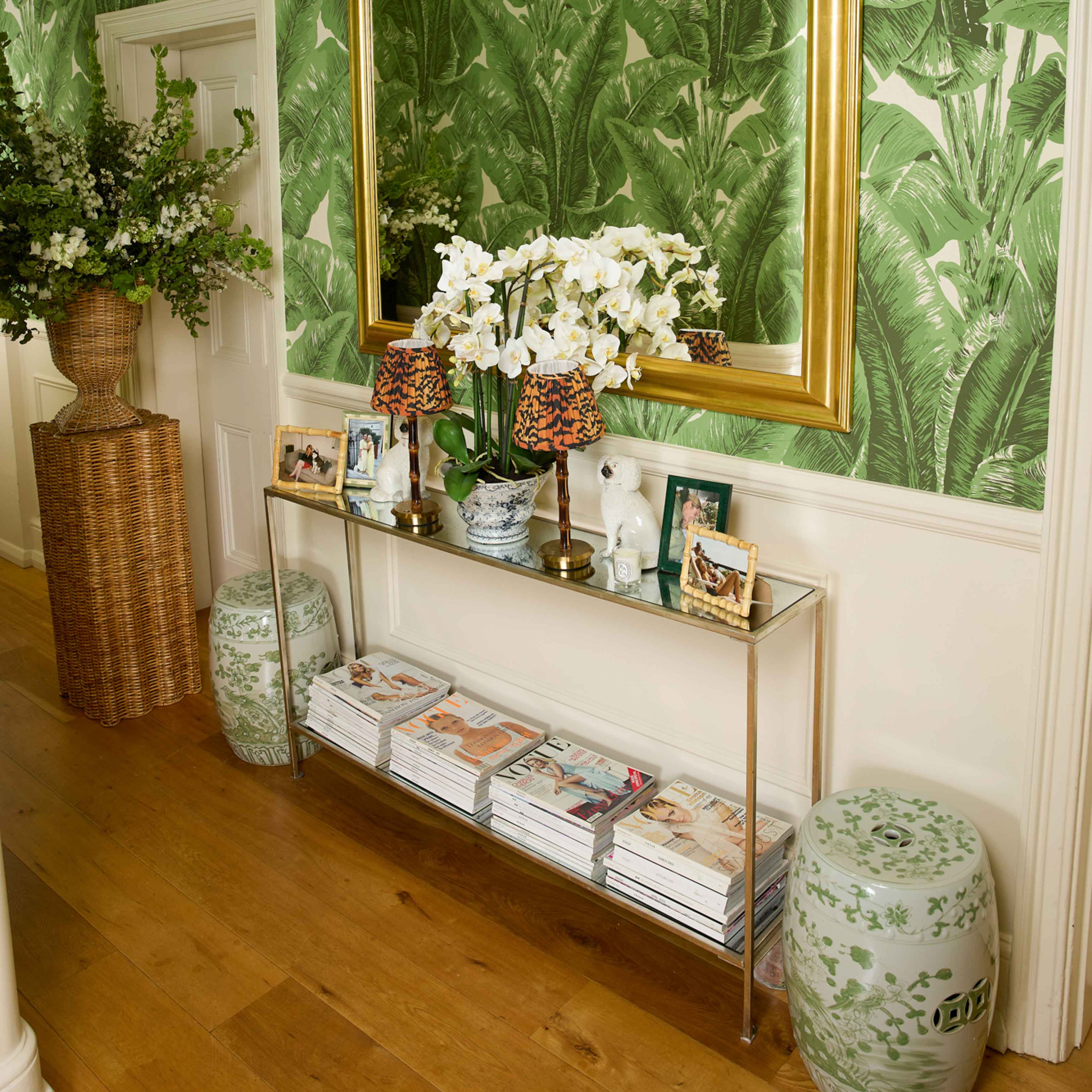 Decorative interior with a console table, mirror, and floral arrangements against a green leafy wallpaper.
