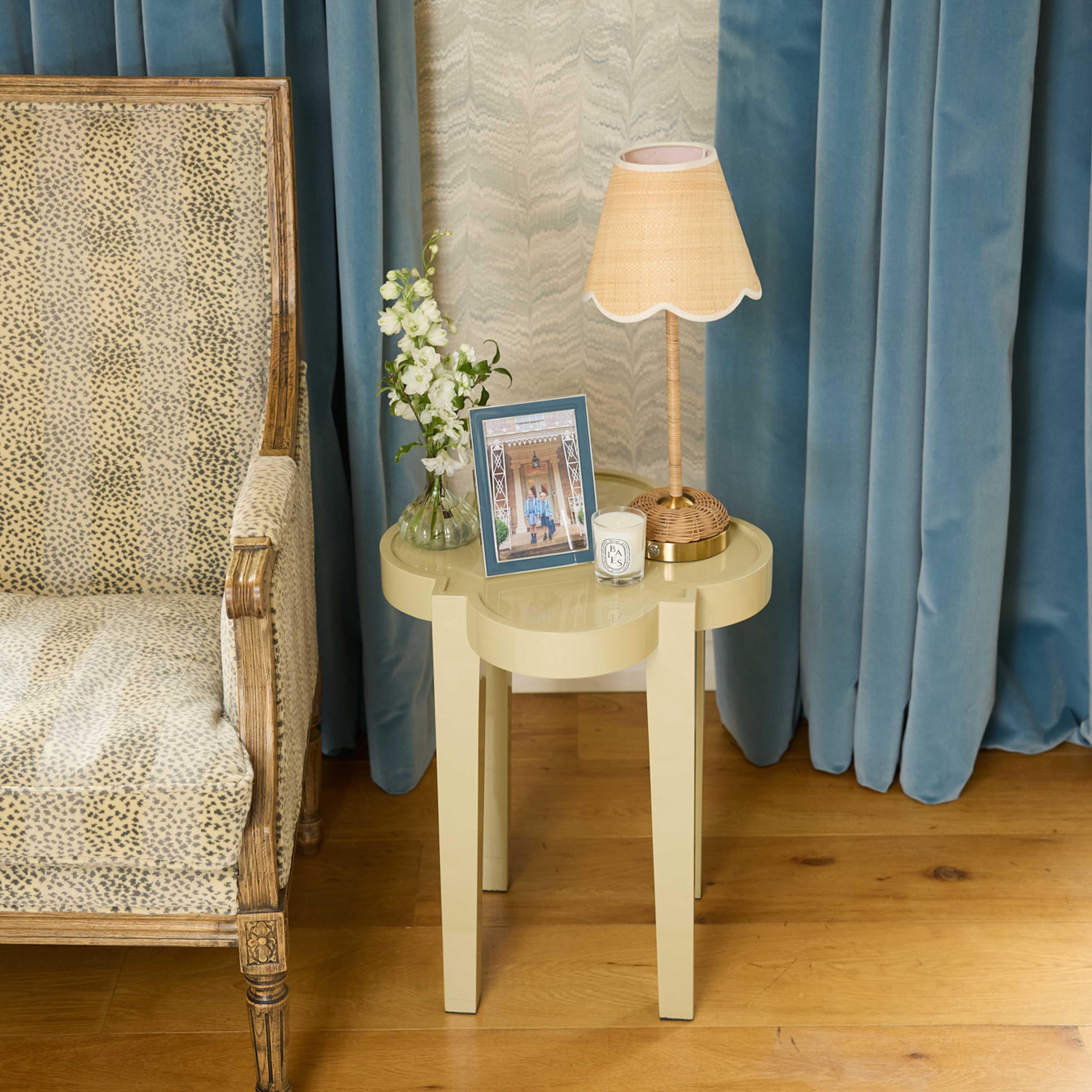 Round wooden side table with lamp, candle, and decorative items next to a patterned chair against blue curtains.
