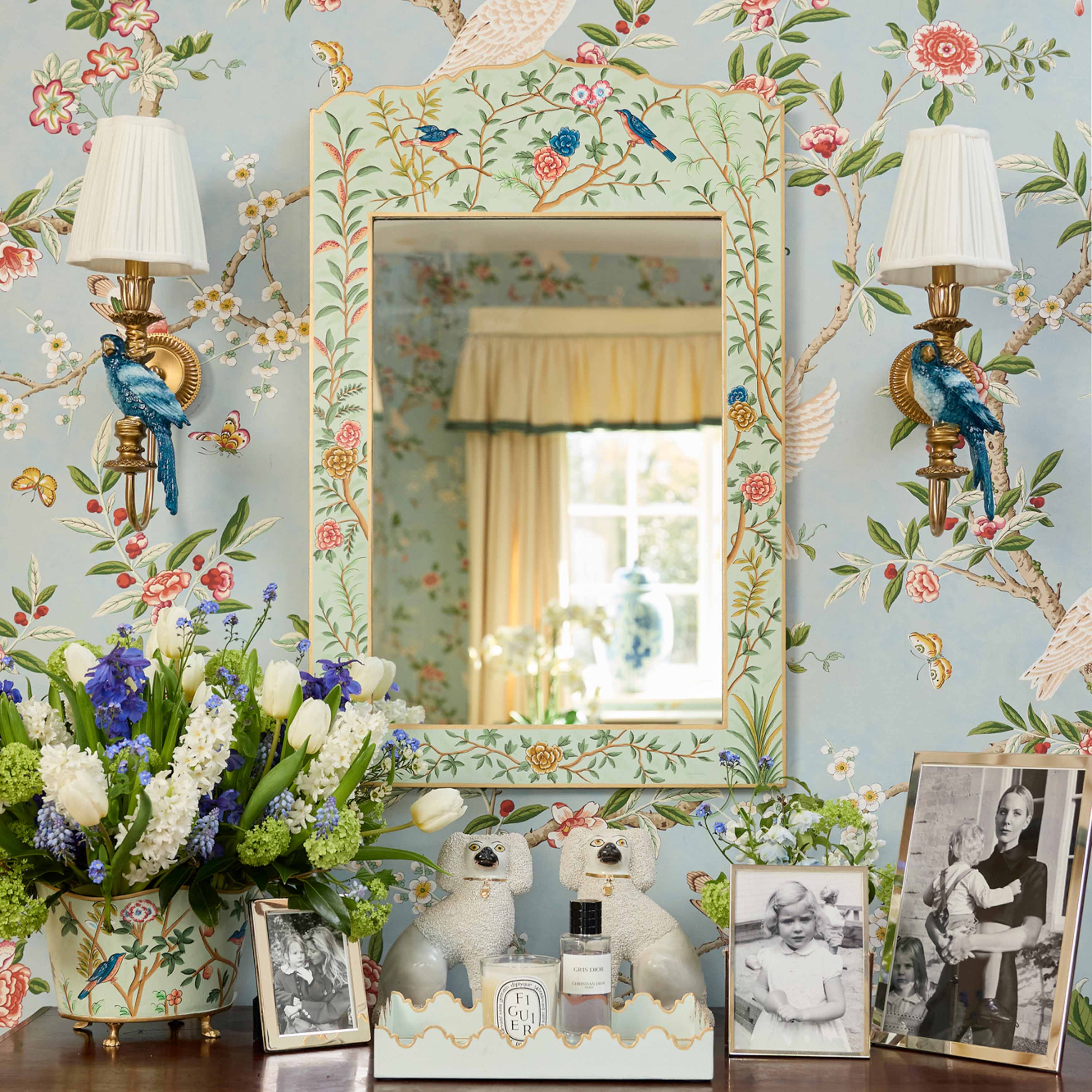 Decorative room interior with floral wallpaper, mirror, lamps, and framed photos.