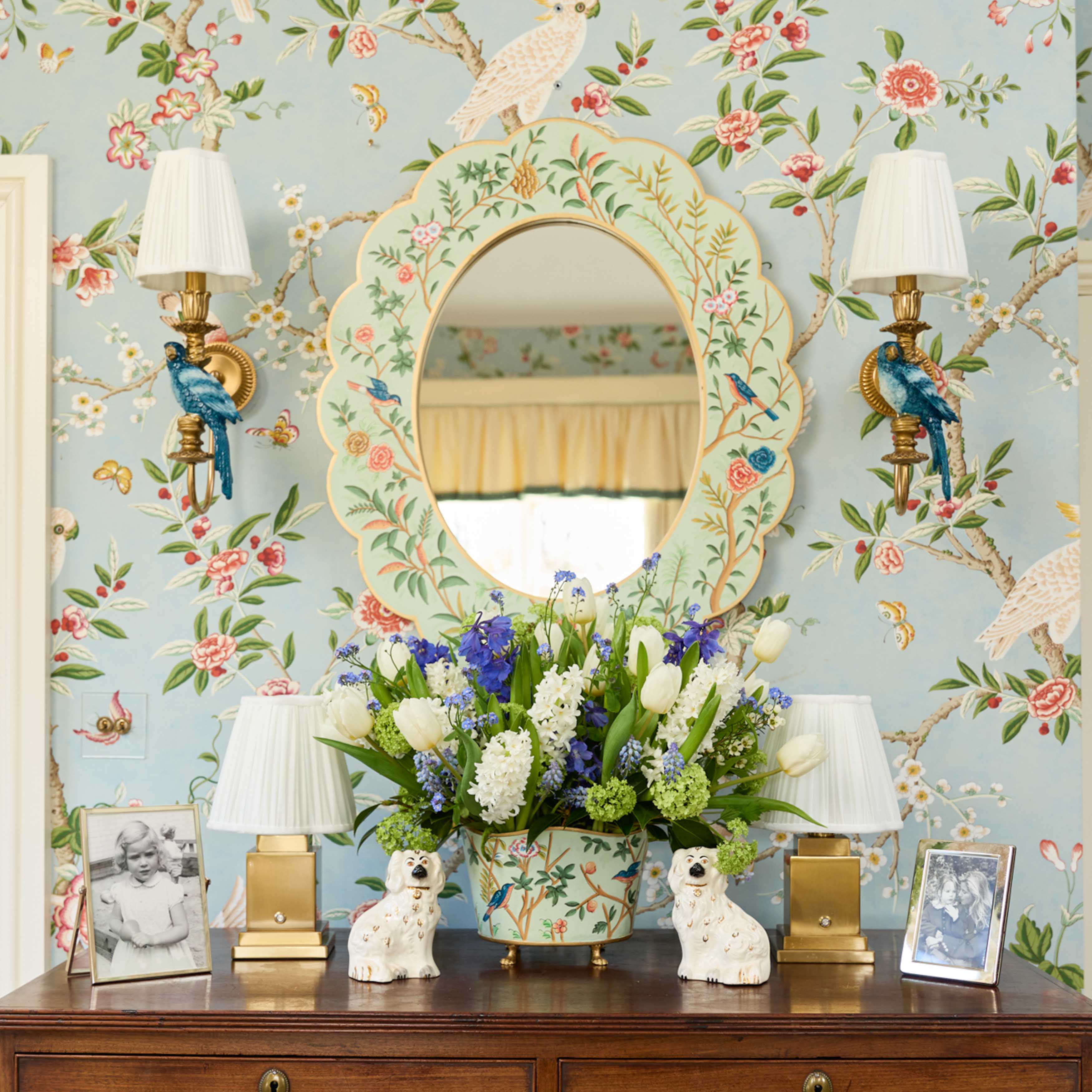 Decorative room interior with floral wallpaper, mirror, lamps, and flowers on a dresser.