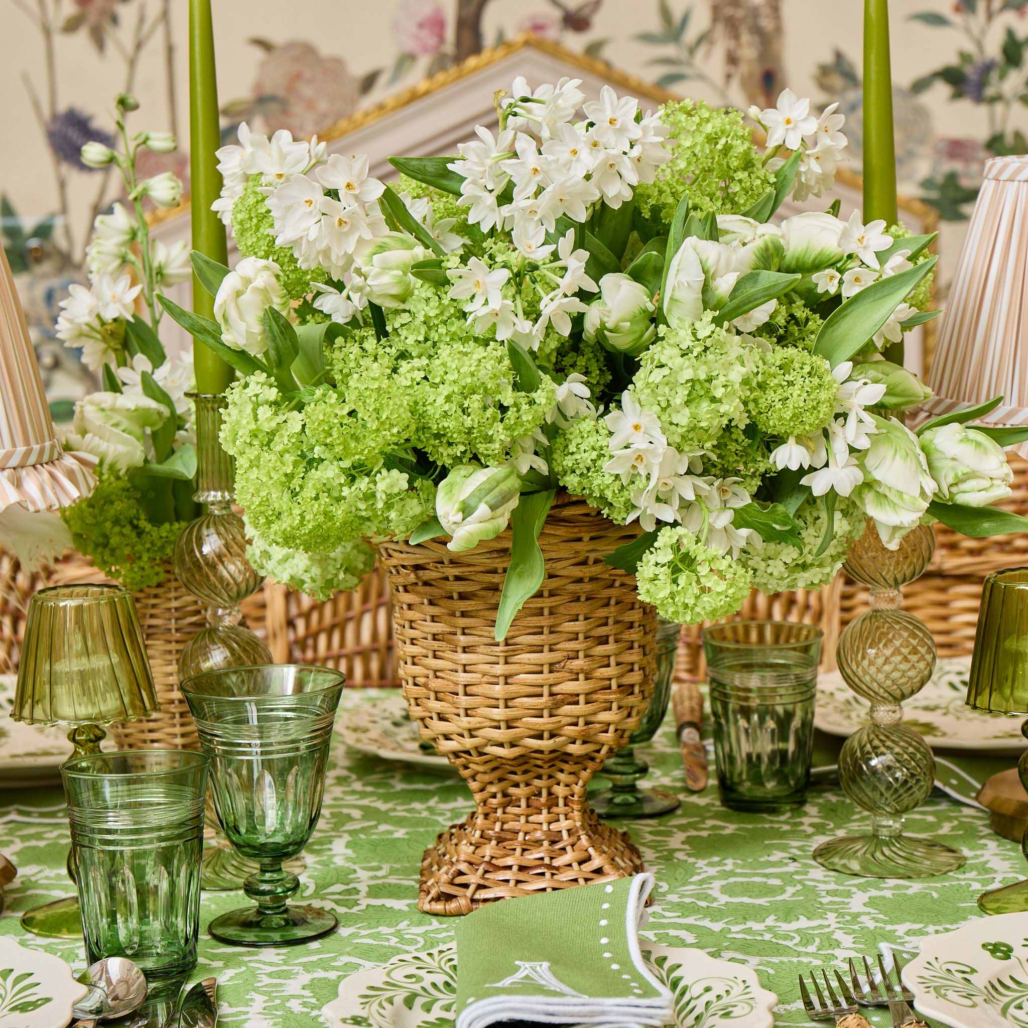 Decorative table setting with a floral arrangement in a woven basket, green glasses, and a patterned tablecloth.