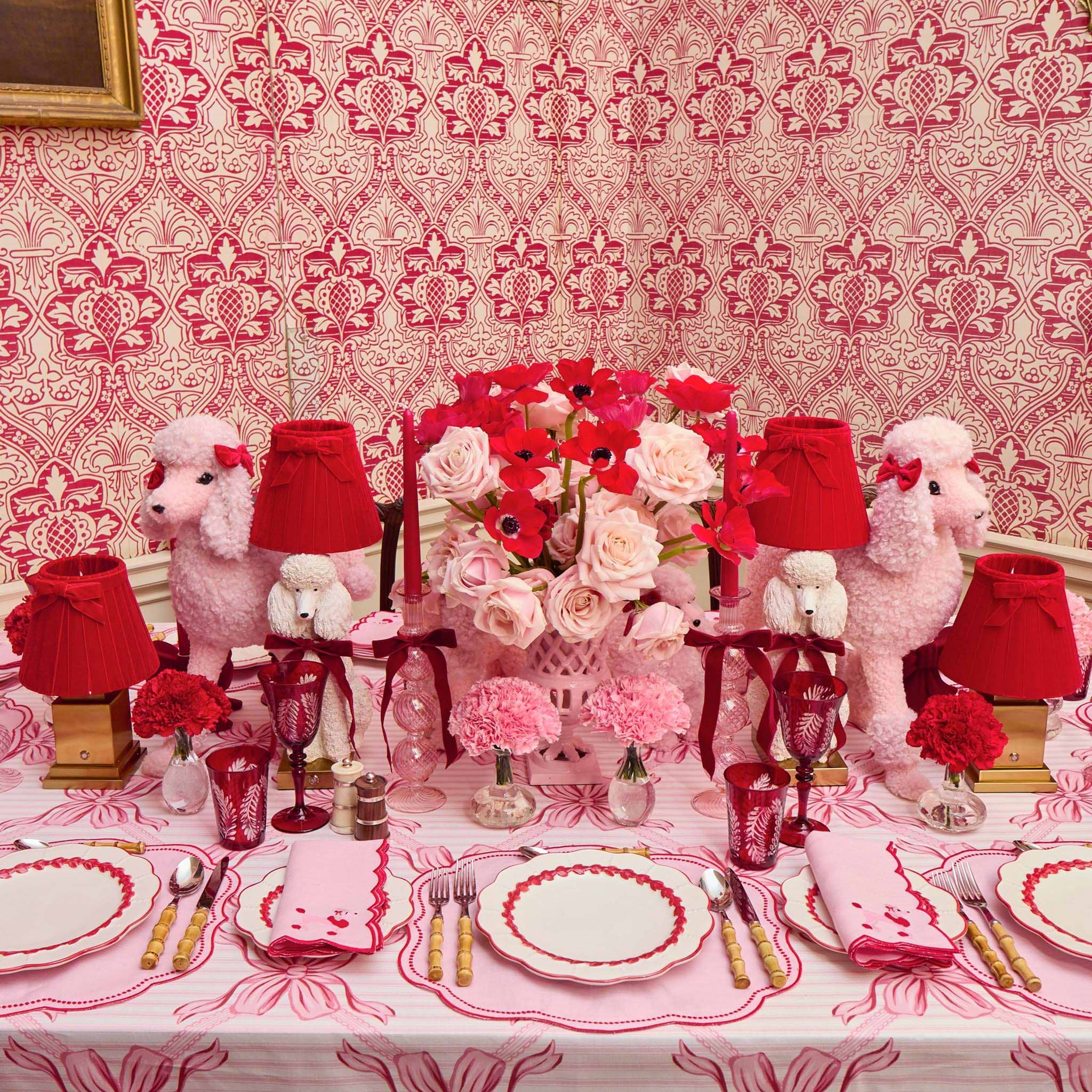 Decorative table setting with poodle figures, red and white tablecloth, and floral arrangements against a patterned wall.