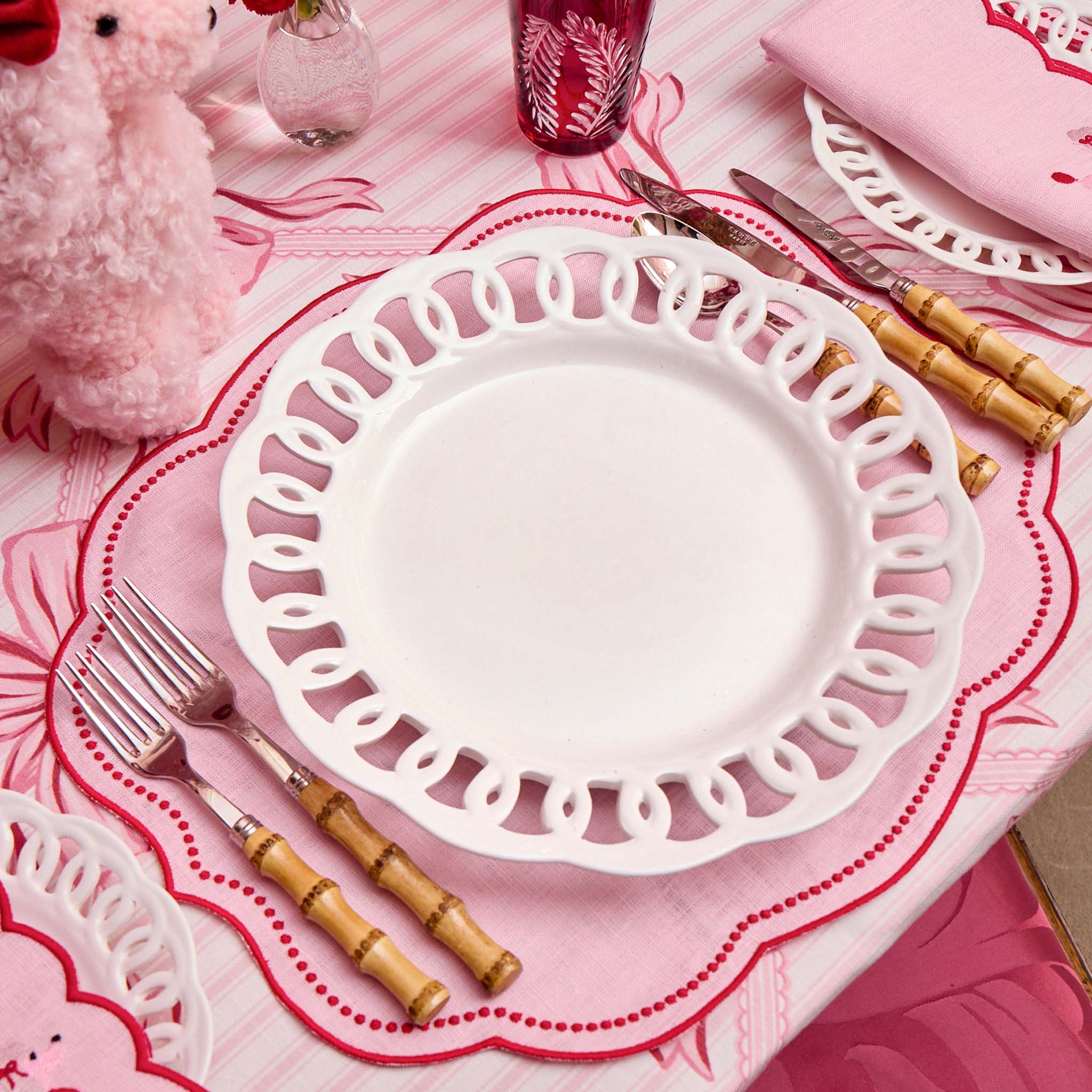 White decorative plate on a pink tablecloth with cutlery and a pink plush toy.