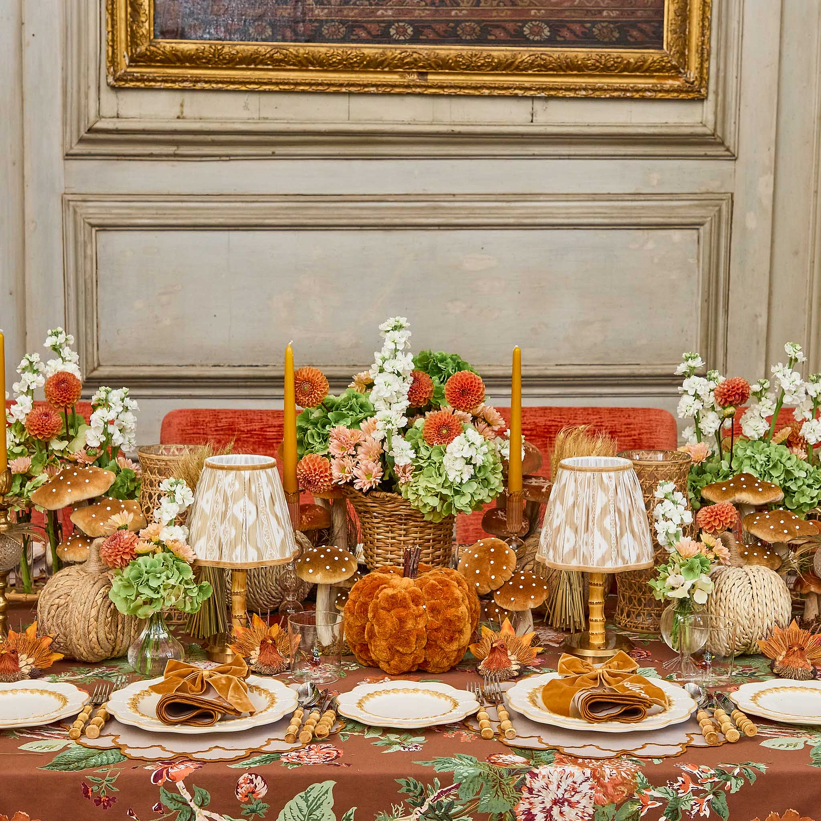 Decorative table setting with pumpkins, bamboo cutlery set, flowers, and lamps in a room with framed artwork on the wall.