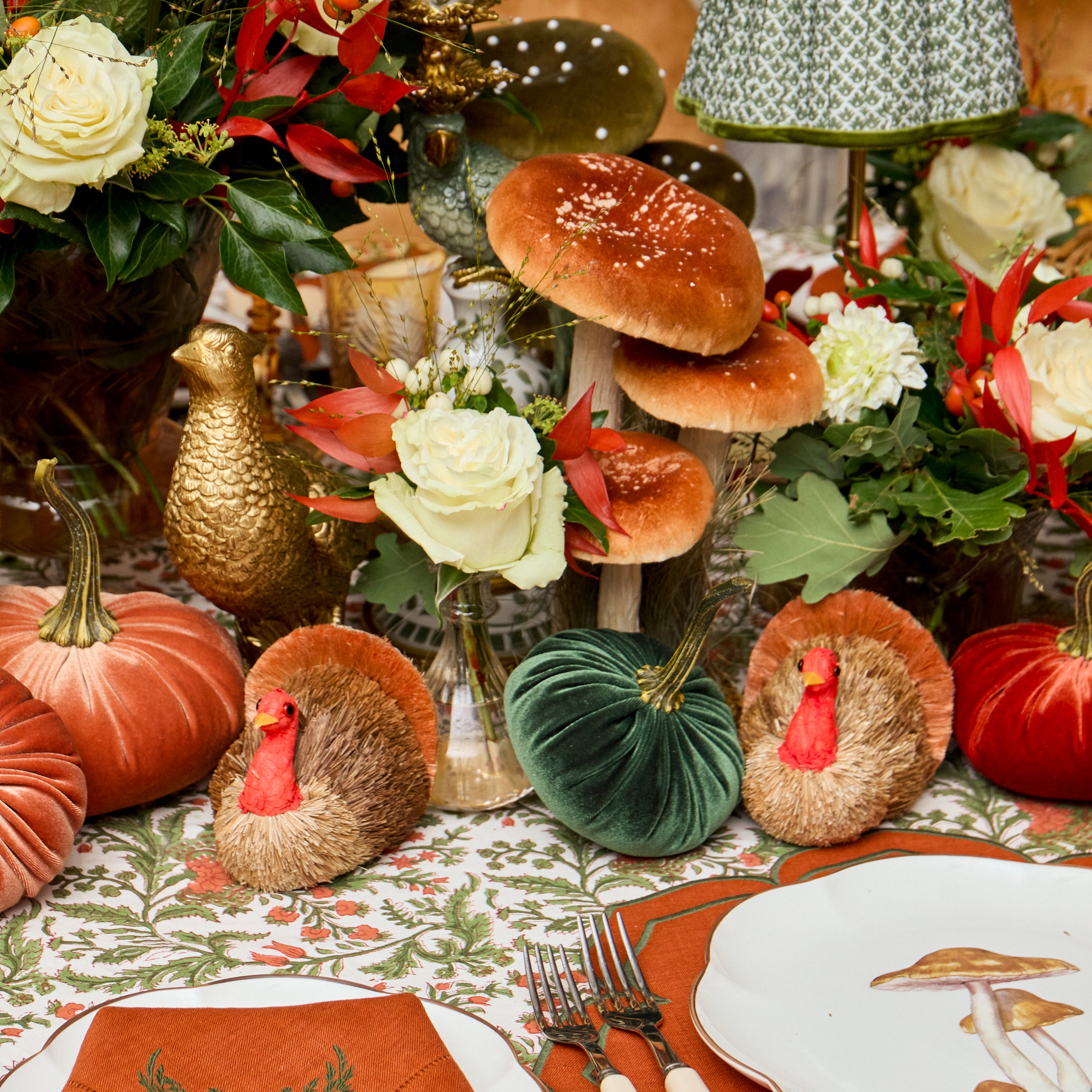 A styled Thanksgiving table centerpiece featuring decorative pumpkins, velvet pumpkins, two turkey-shaped ornaments, large brown mushrooms, white roses, red foliage, and floral table linens.