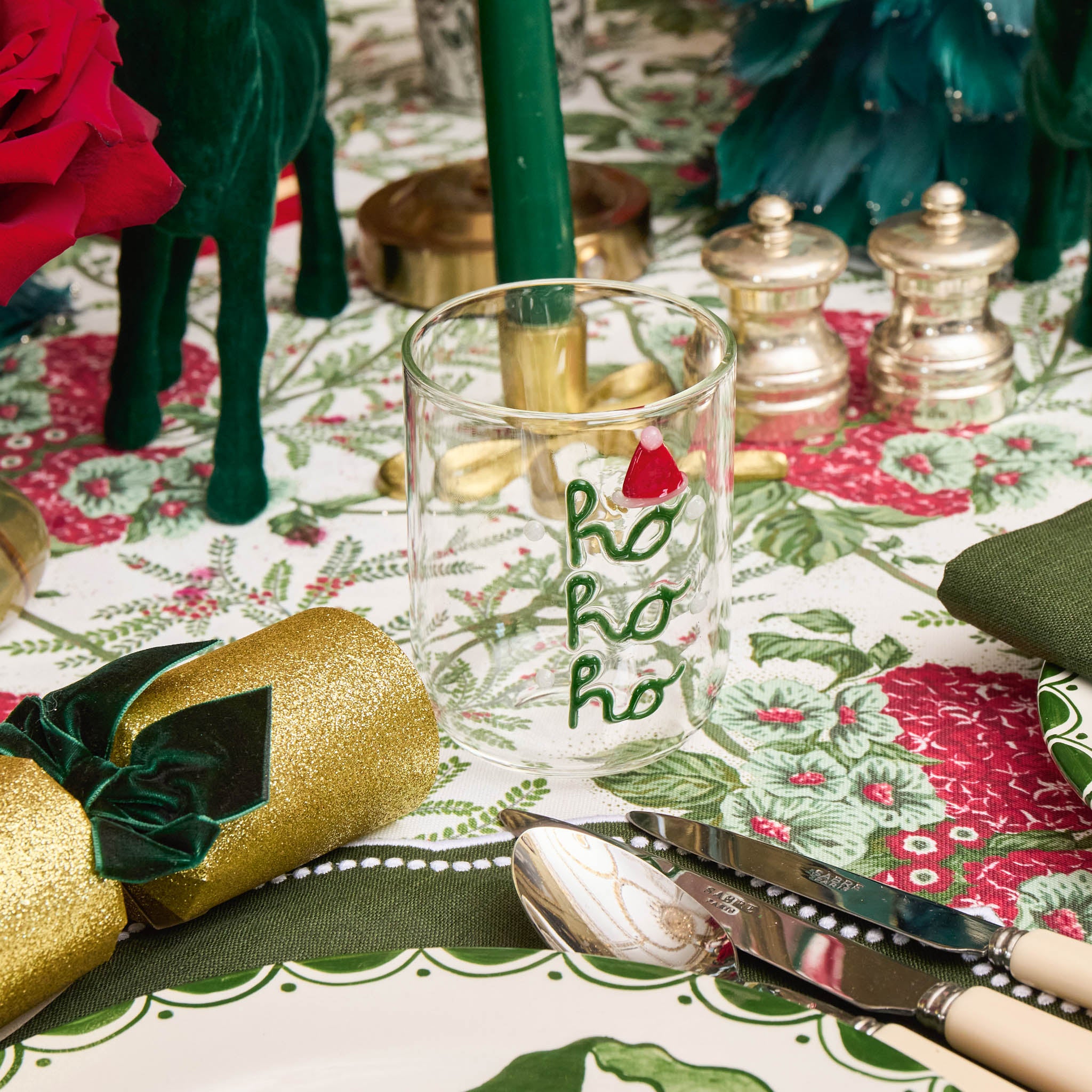 Decorative table setting with a glass, gold cracker, and red roses on a patterned tablecloth.