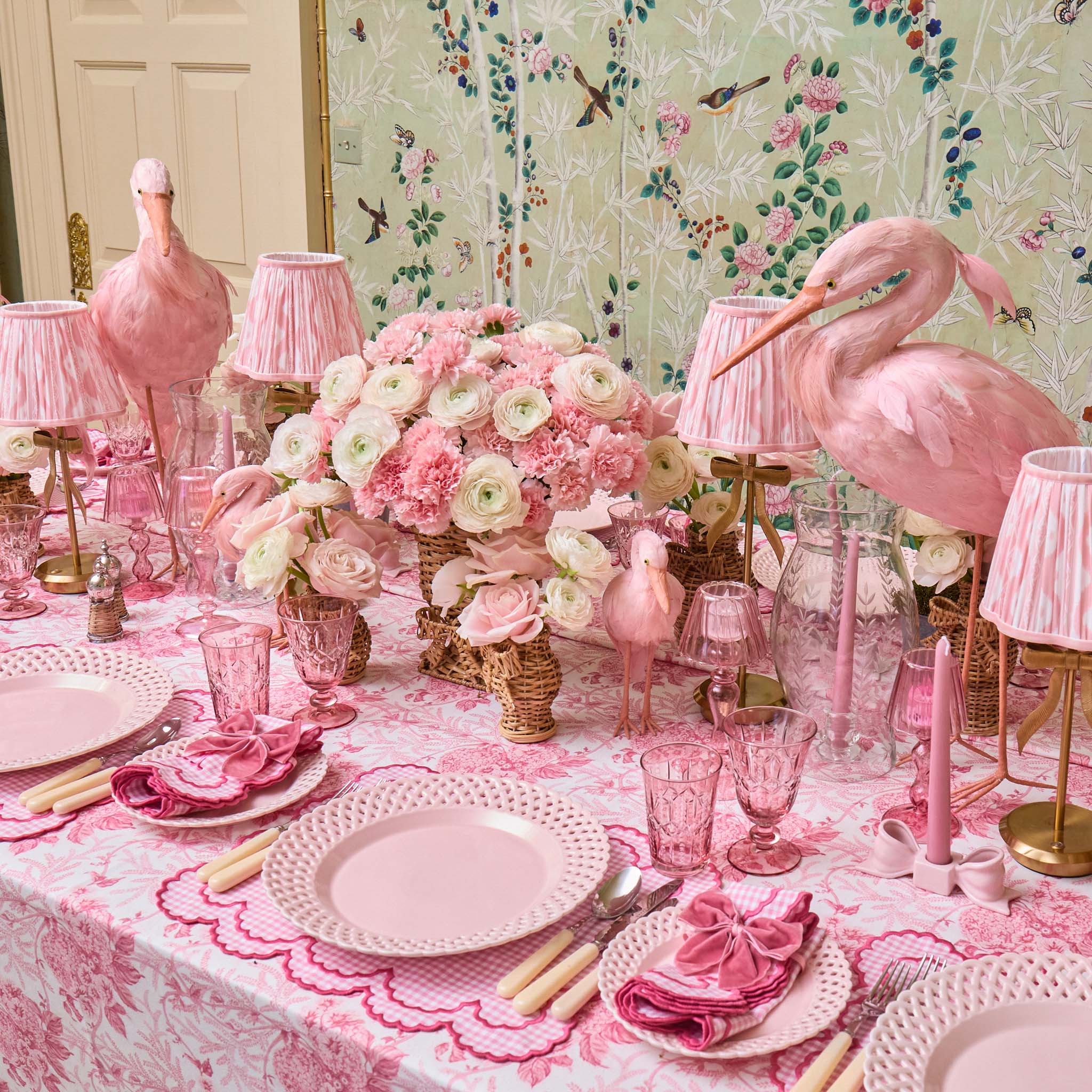 Pink flamingo-themed table setting with floral arrangements and decorative lamps against a floral wallpaper background.