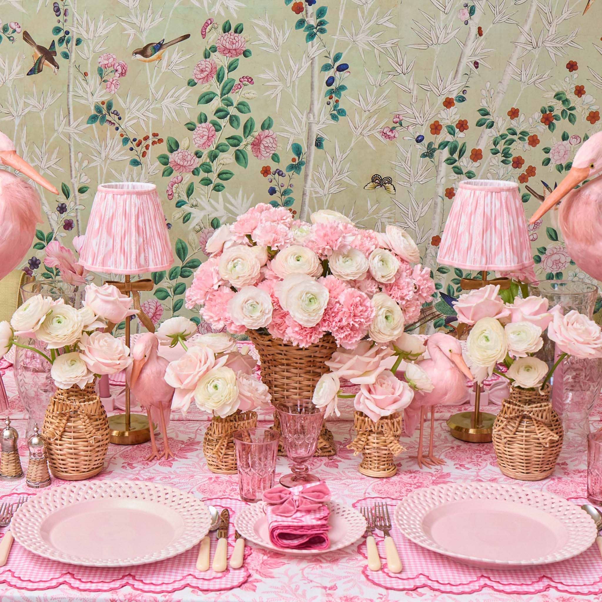 Decorative table setting with pink and white flowers, lamps, and plates against a floral wallpaper background.
