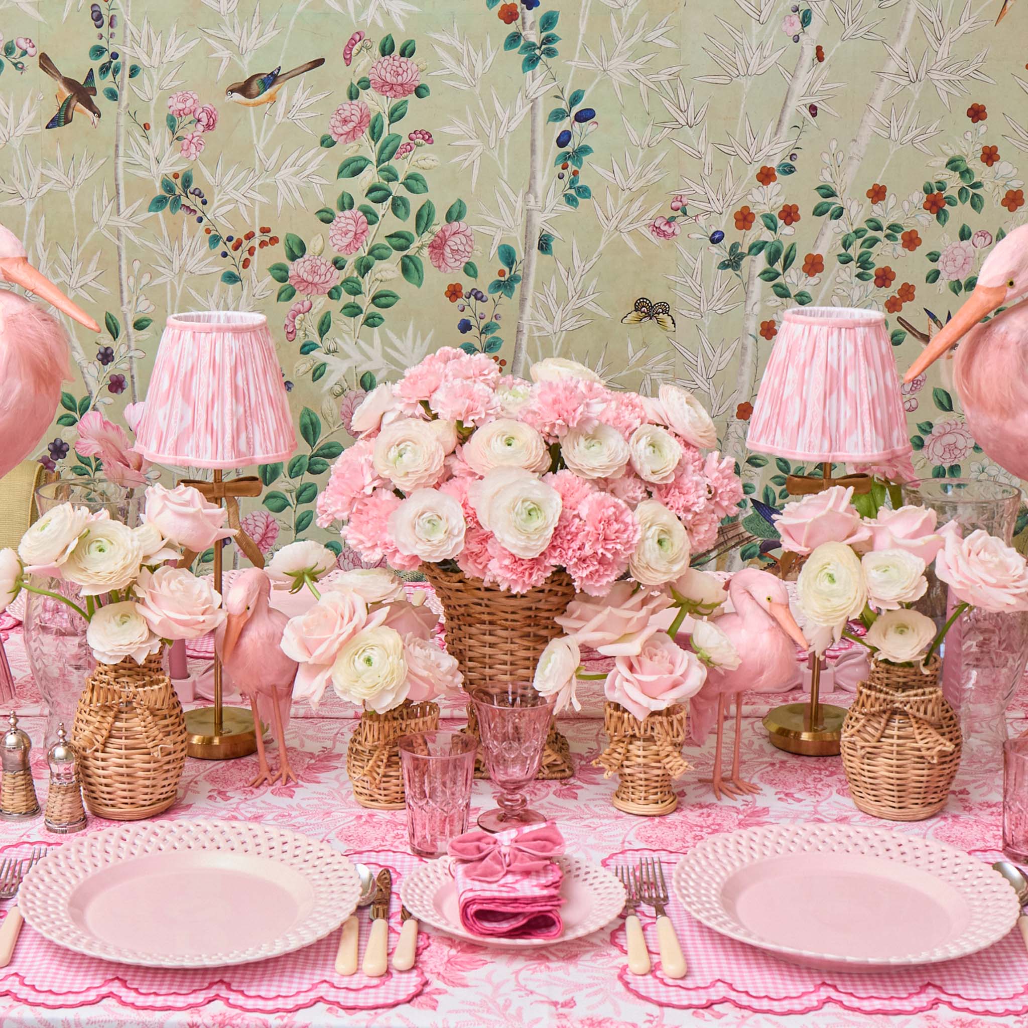 Decorative table setting with pink and white flowers, lamps, and plates against a floral wallpaper background.