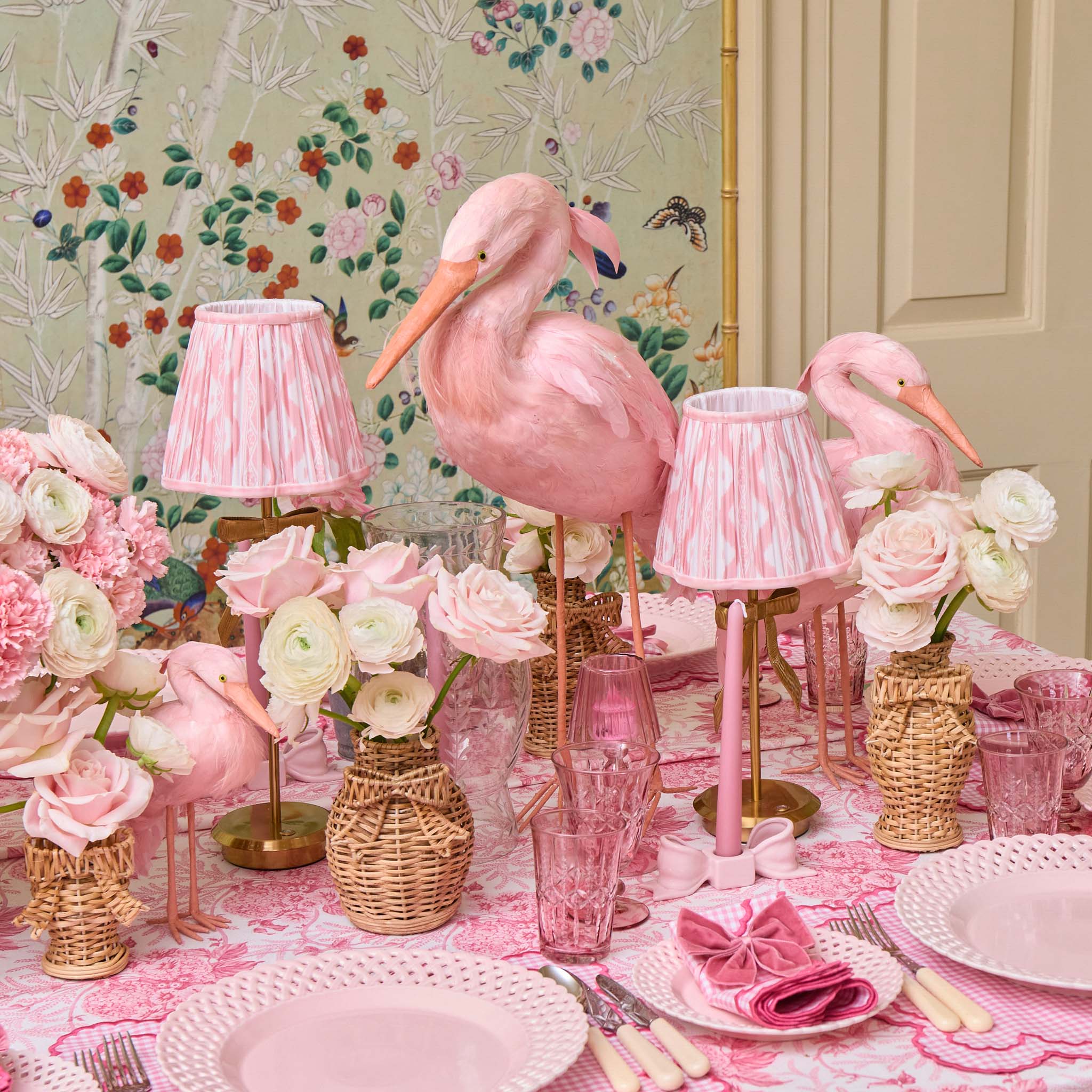 Decorative table setting with pink flamingo lamps and floral arrangements against a floral wallpapered wall.