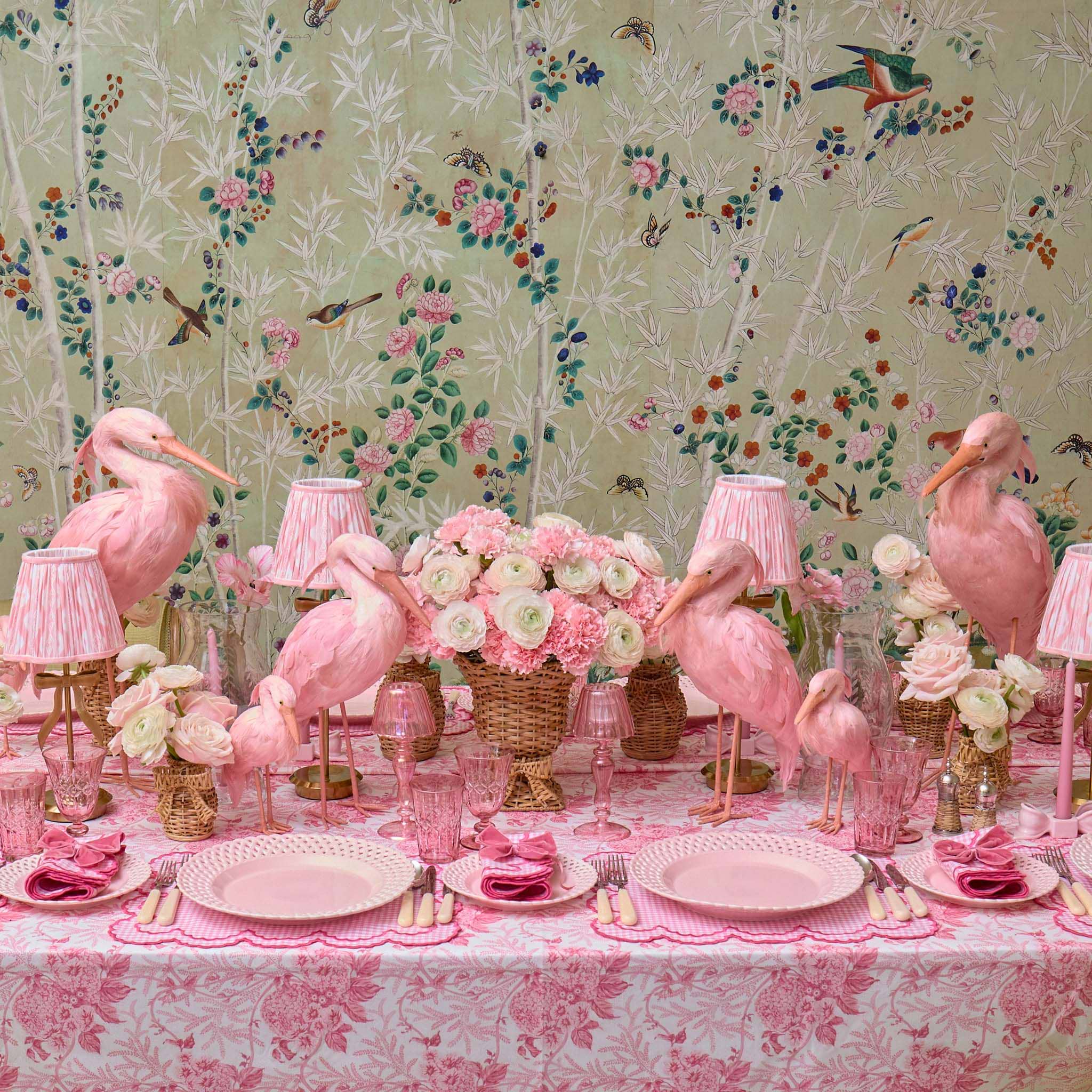 Decorative table setting with pink flamingo figurines, floral arrangements, and lamps against a floral wallpaper background.
